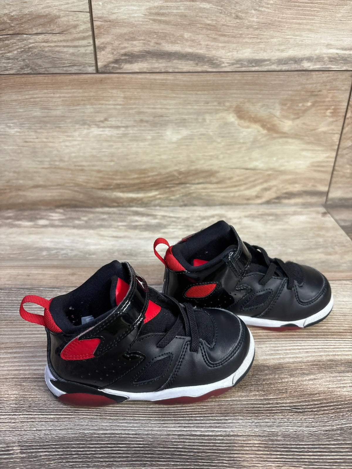Angled view of Air Jordan Flight Club 91 ‘Bred’ toddler sneakers, black with red accents and Velcro strap on wood floor