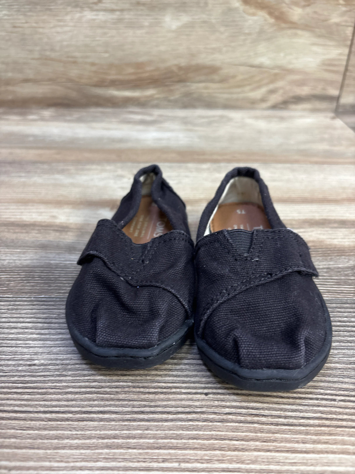 Front view of Toms Tiny Alpargata toddler shoes showcasing black canvas upper and rubber sole on wood background