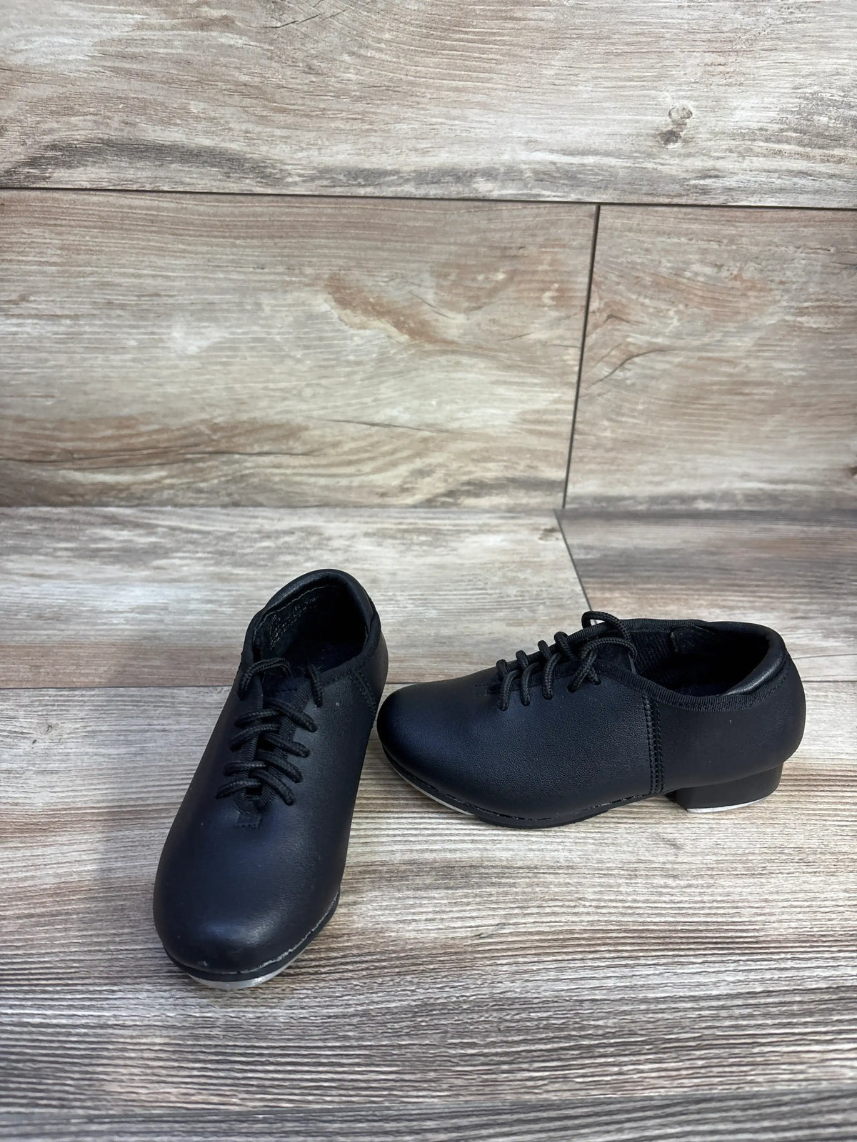 Angled front view of children’s black Theatricals tap shoes showcasing laces, low heel, and metal taps with wood-look background