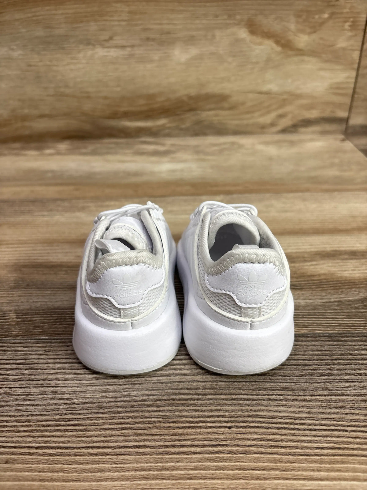 Rear view of Adidas X_PLR sneakers showing white uppers, padded collars, and thick soles on a wood-grain surface