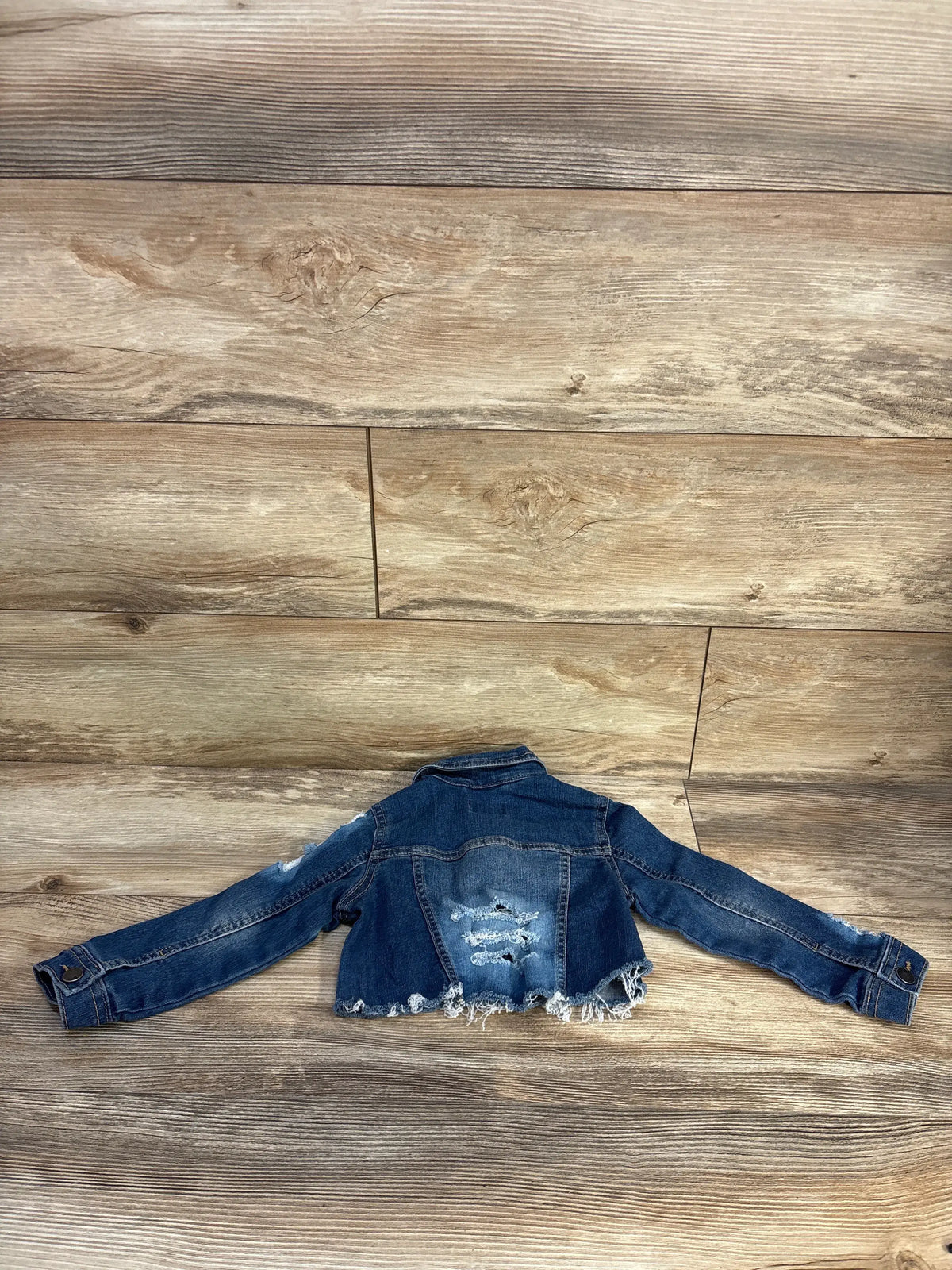 Back view of Children’s Place blue distressed denim jacket in size 2T showcasing frayed hem and back distressing on a wood floor