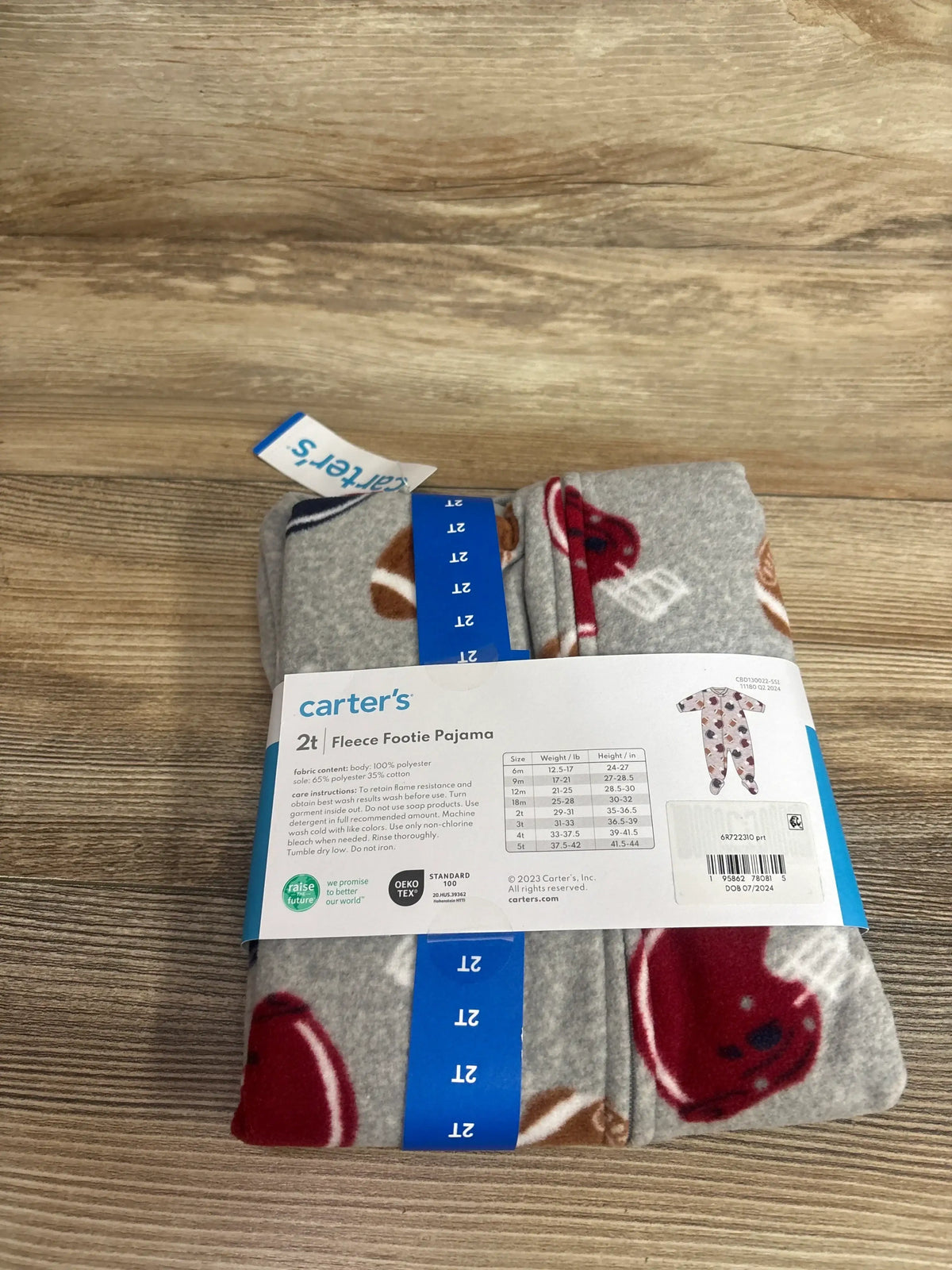 Top-down of folded Carter’s grey fleece footie pajama with football and helmet print, size 2T visible on blue band on wood background