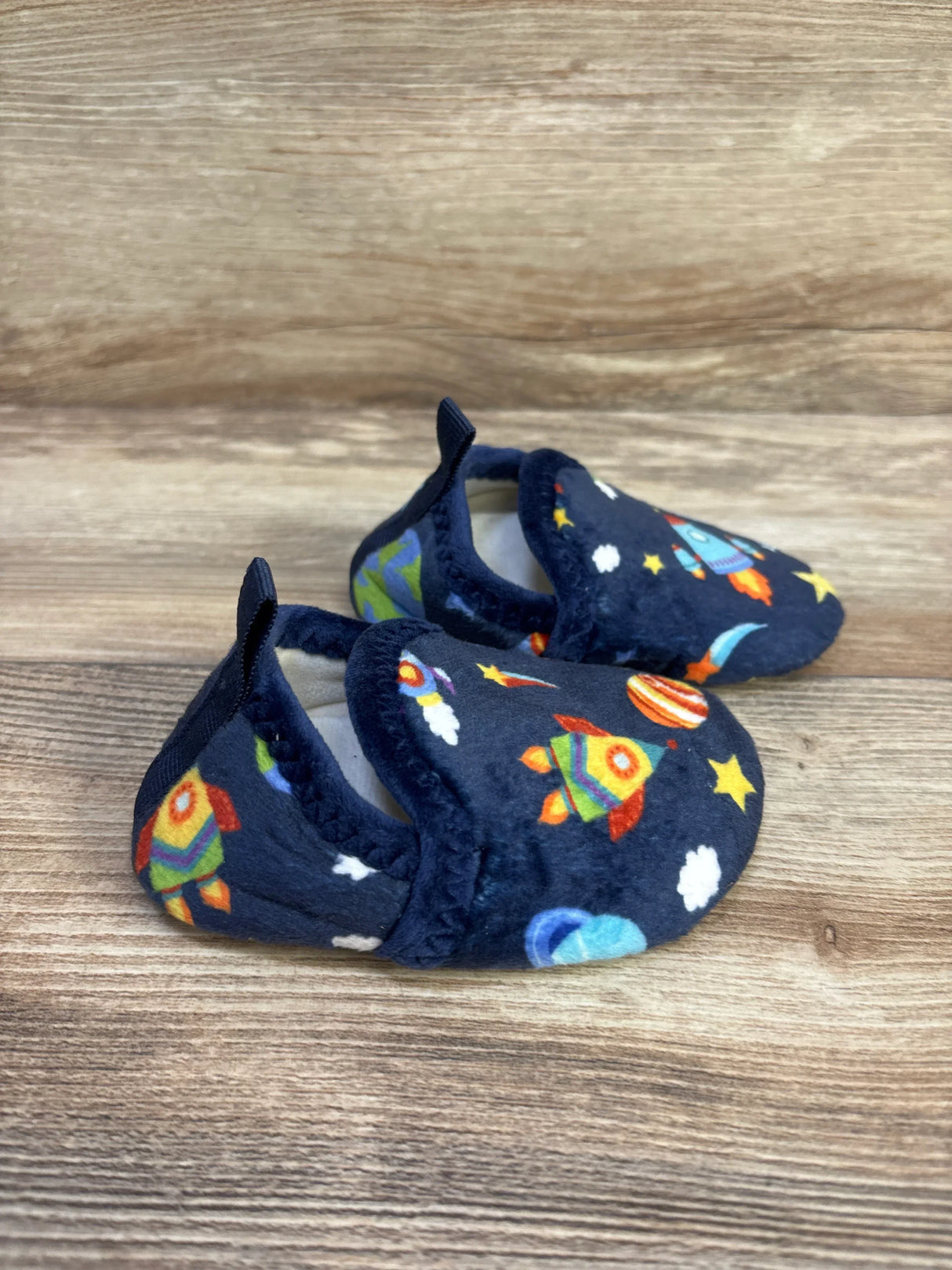 Three-quarter view of navy baby slip-on slippers showing colorful rocket and planet print, plush lining and heel pull tab on wood surface