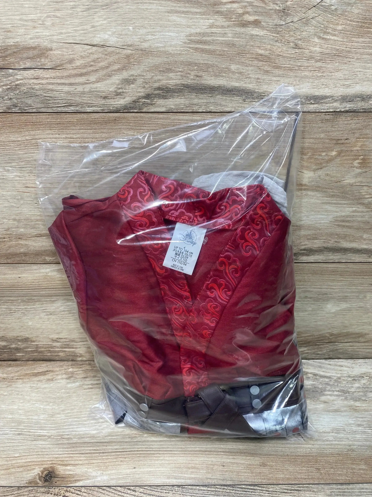 Front view of folded Disney 3-piece Mulan toddler costume with embroidered red collar and brown belt in clear bag on wood background