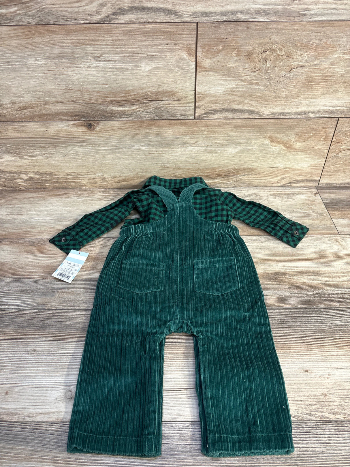 Flat lay of green corduroy baby overalls and green plaid shirt showing front pockets and cross-back straps on wood background