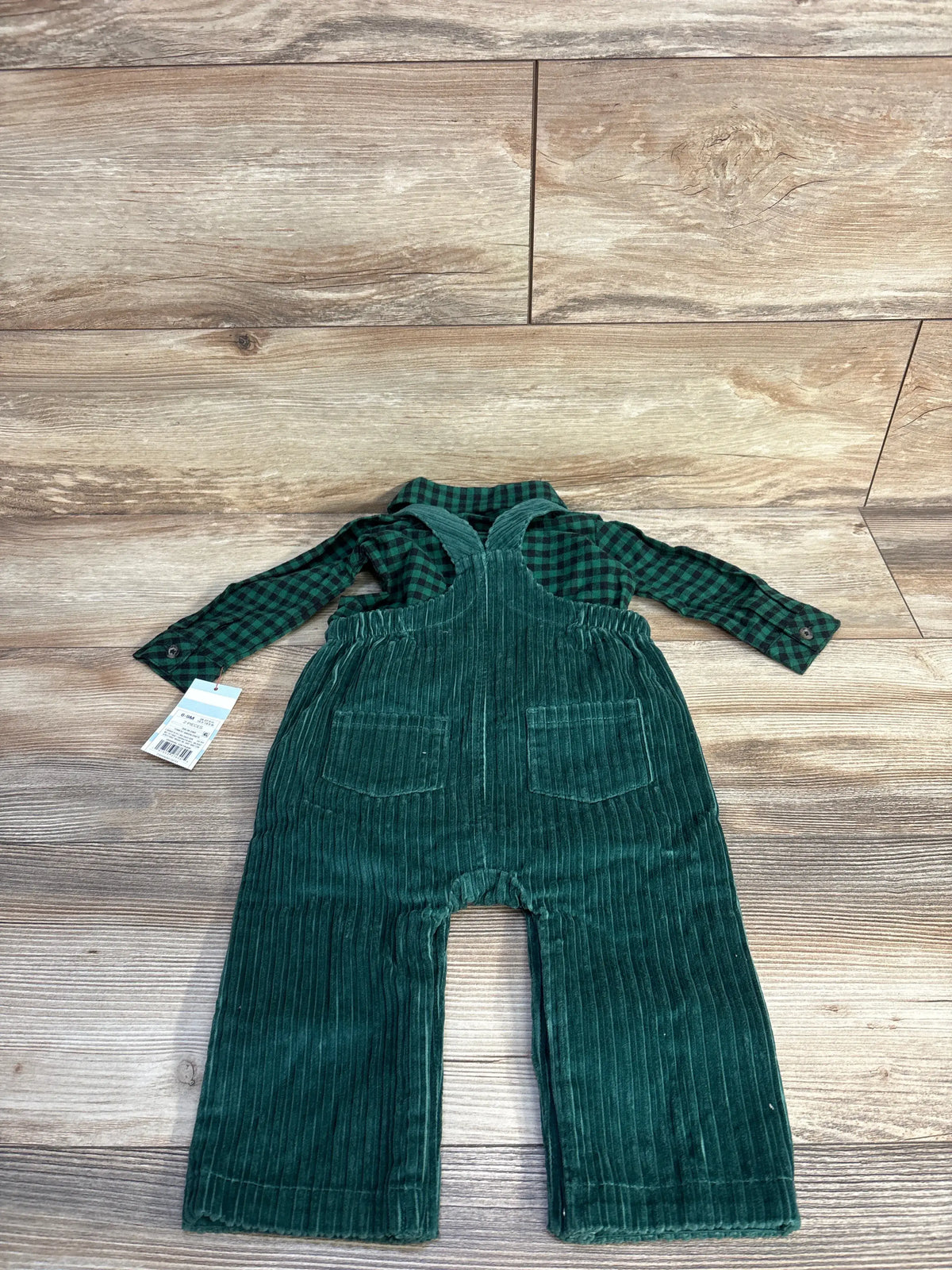 Flat lay of green corduroy baby overalls and green plaid shirt showing front pockets and cross-back straps on wood background