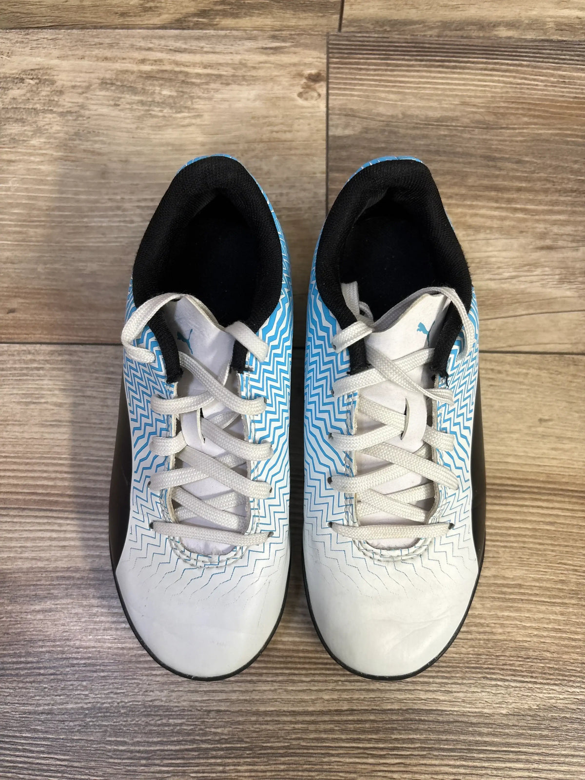 Top view of Puma Rapido II soccer cleats size 12C showing white upper with blue zigzag pattern and black heel on wood floor