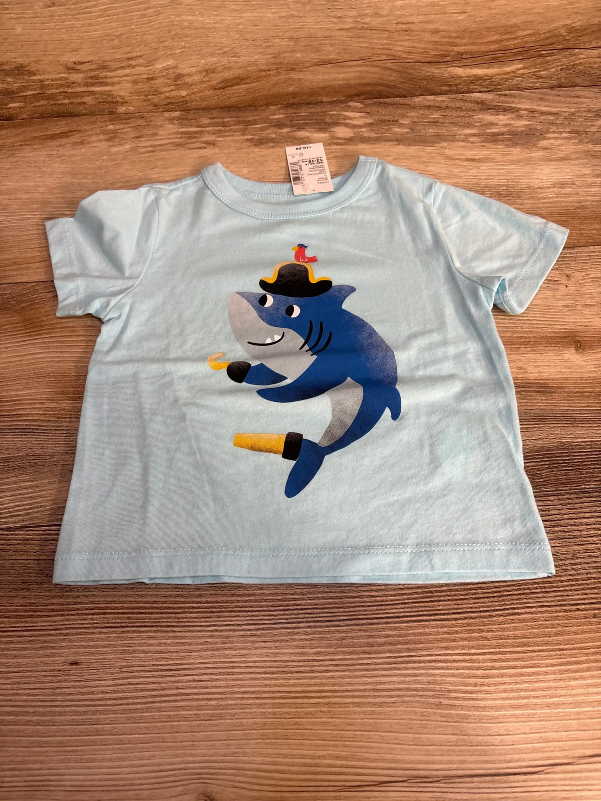 Front flat-lay of light blue toddler short-sleeve shirt showcasing pirate shark graphic with size tag visible on wood background
