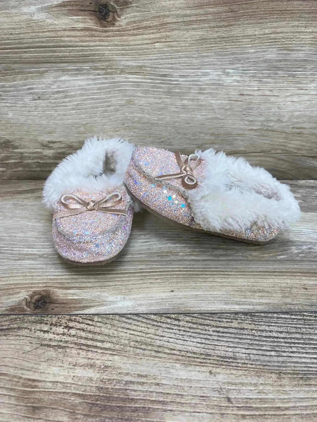 Three-quarter view of Cat & Jack Medora toddler slippers (size 6C) showcasing iridescent pink glitter exterior, faux-fur lining and bows on wooden surface