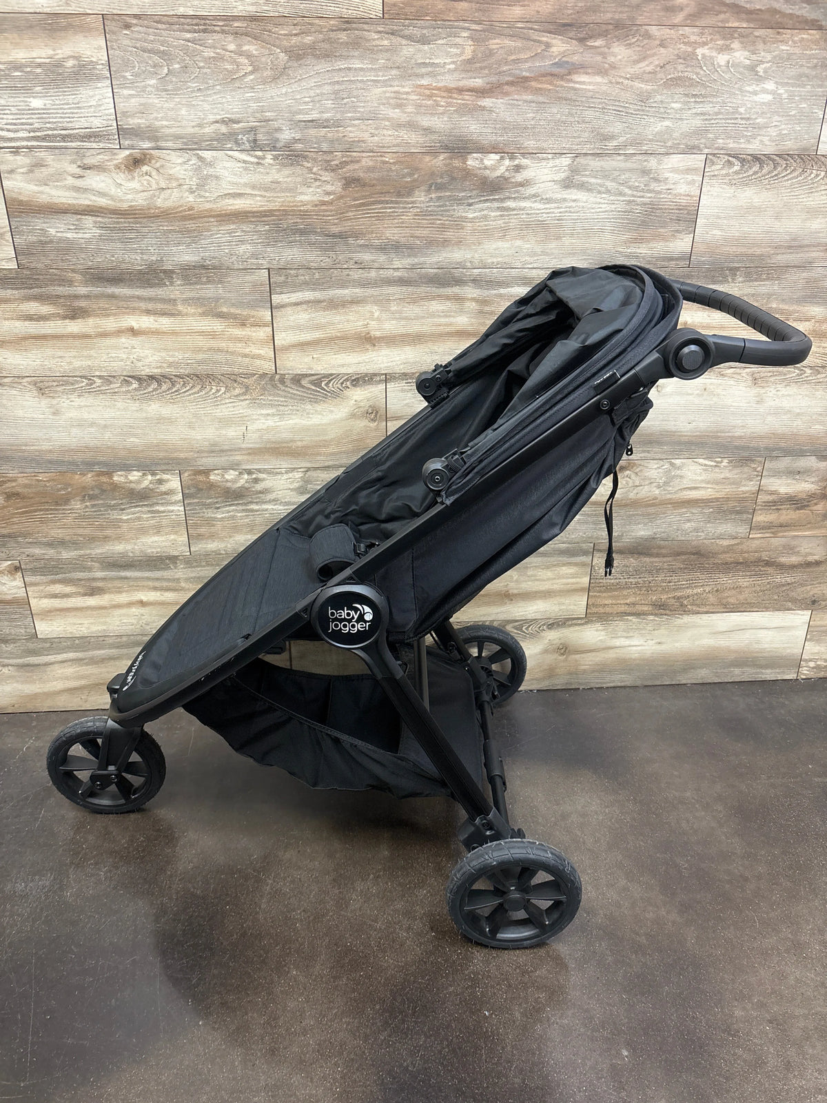 Angled view of Baby Jogger City Mini GT2 All Terrain Stroller in black showcasing three wheels and storage basket against wood paneling
