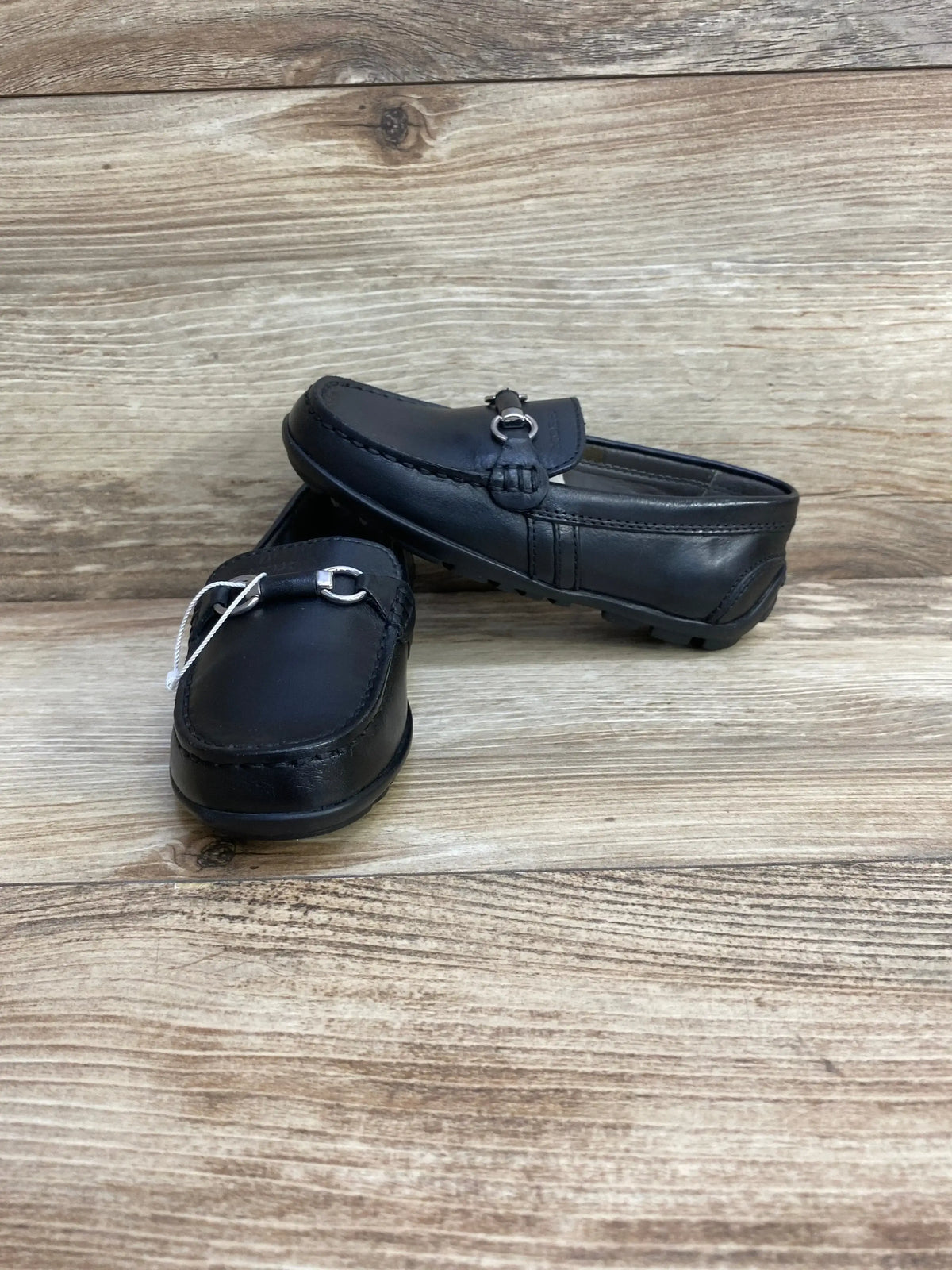 Front three-quarter view of GEOX black leather loafers size 10C showing silver chain detail on wood background