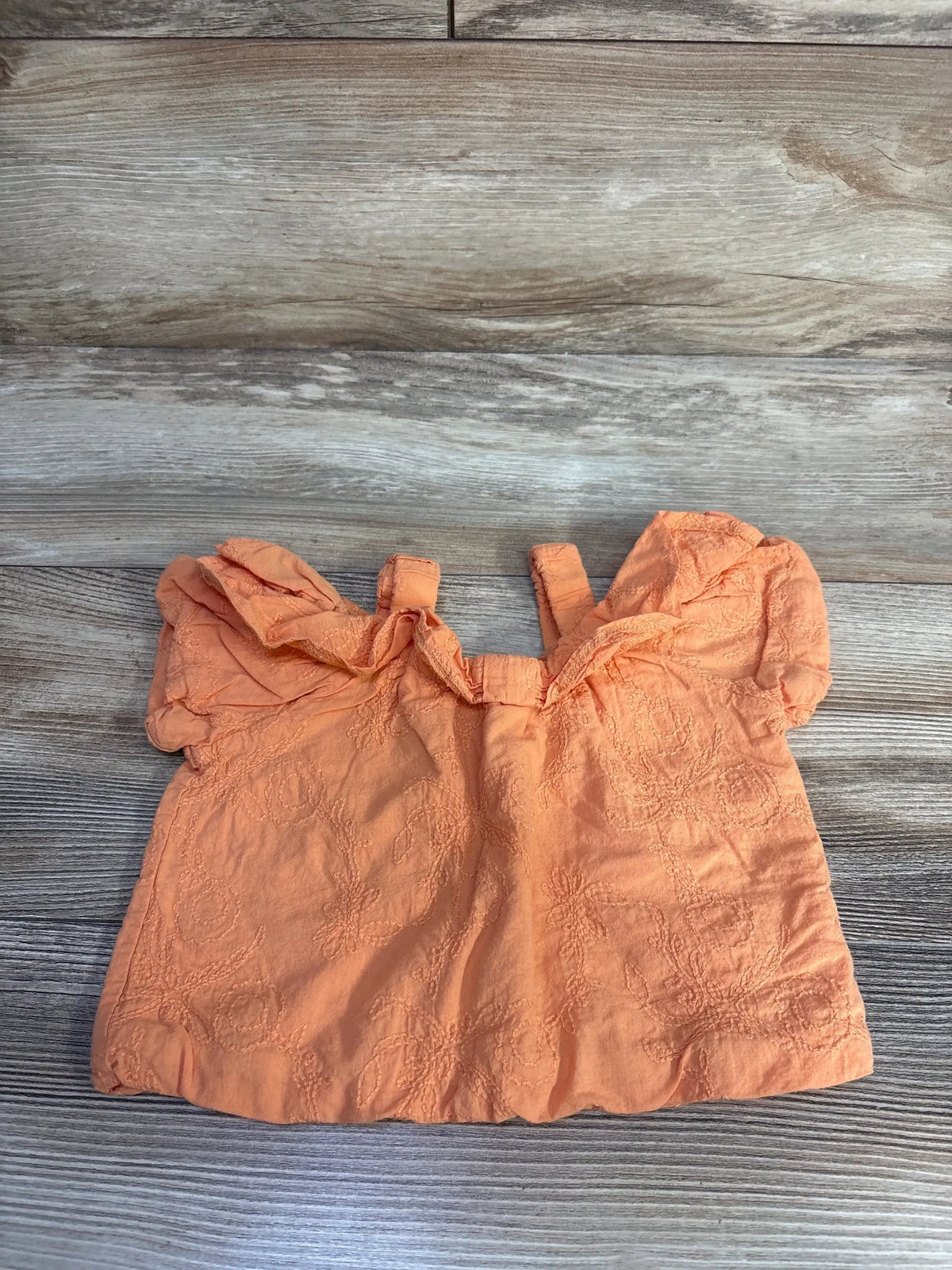 Top-down view of orange Petit Main cold-shoulder baby top showcasing floral embroidery and ruffled sleeves on wood background