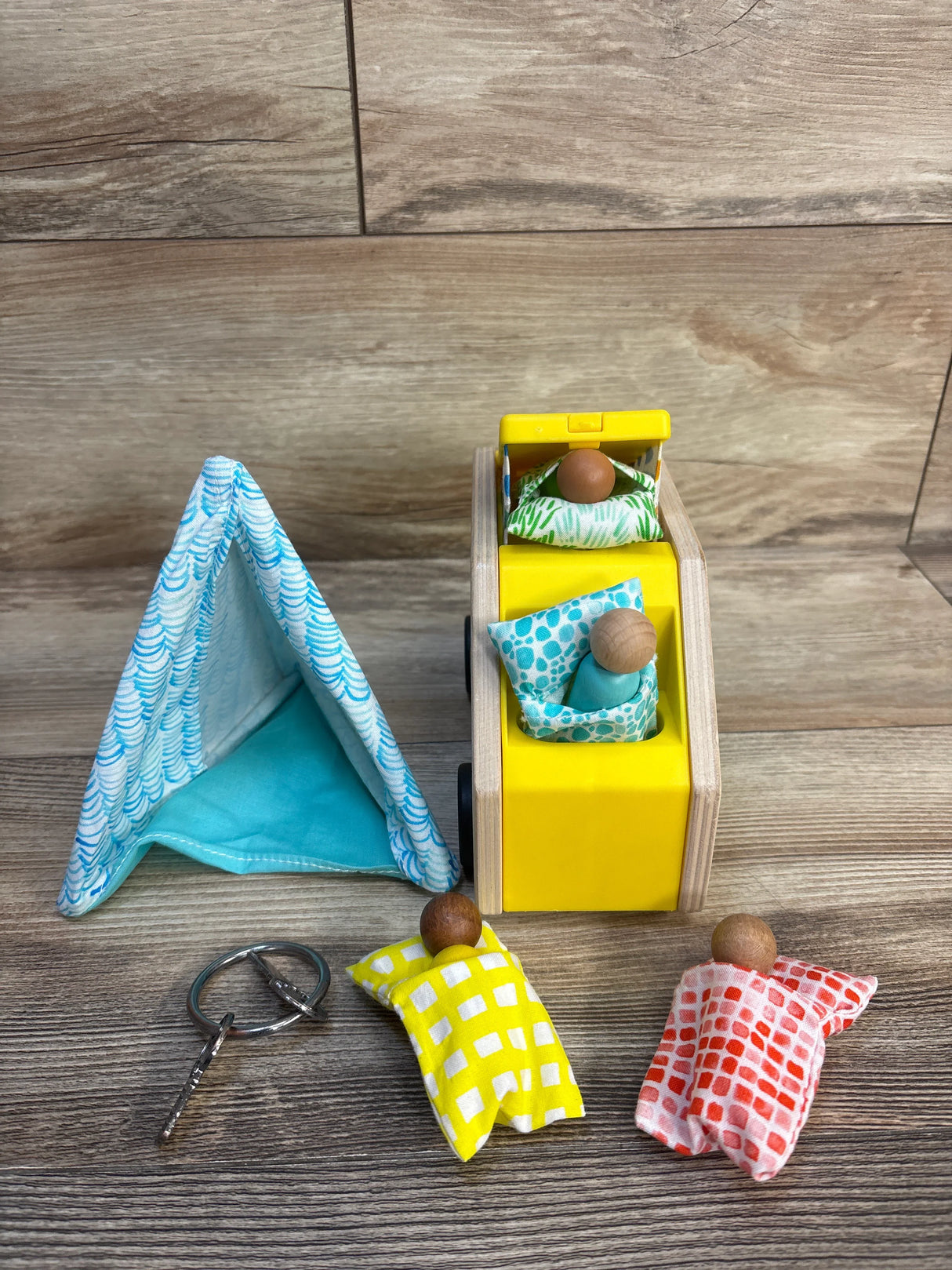 Wooden toy camper set with fabric tent, mini dolls, and colorful sleeping bags on wood floor.