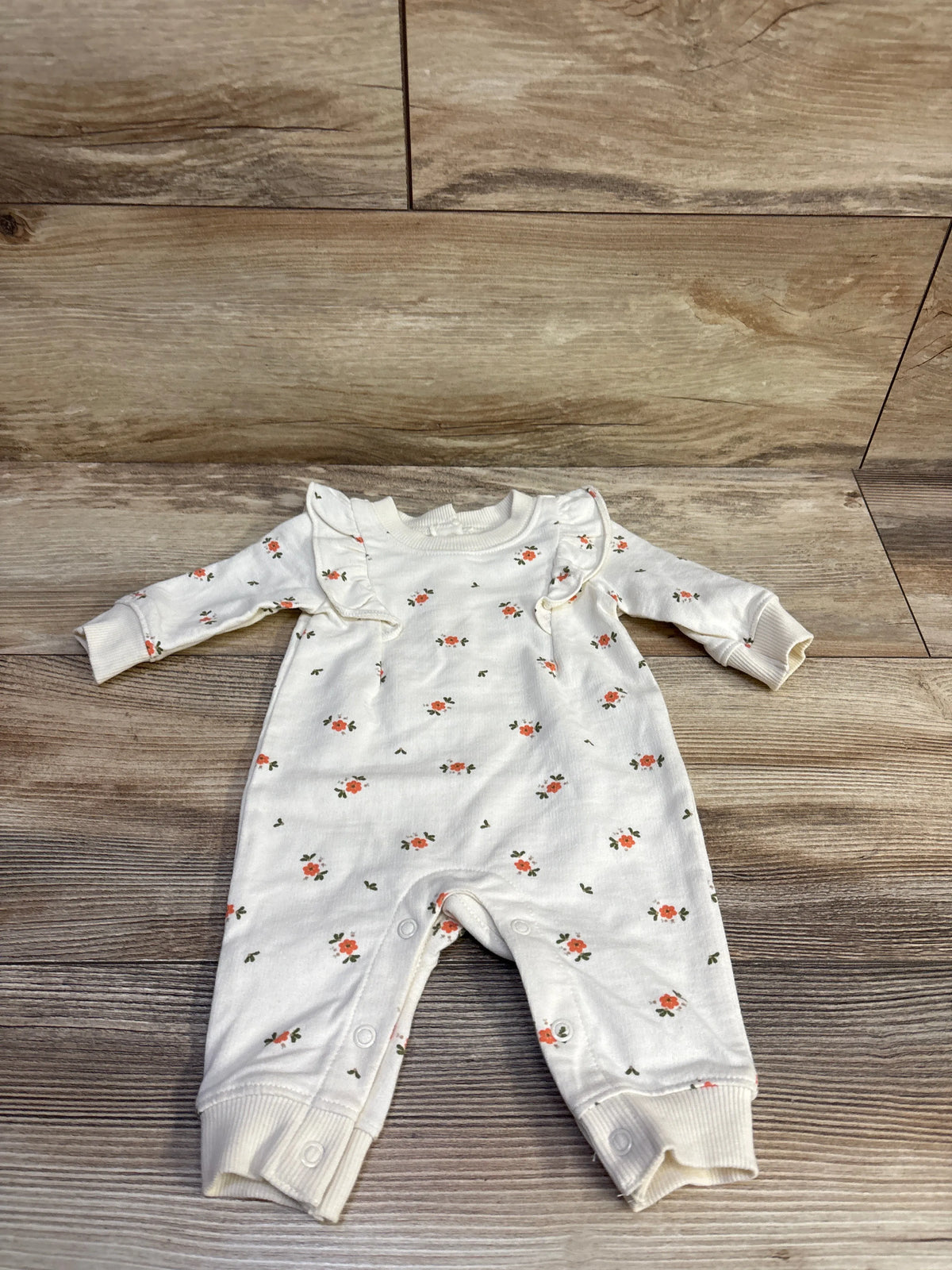 Front view of Carter’s ivory coverall with small pink floral print, ruffle trim, and snap leg closures on wood