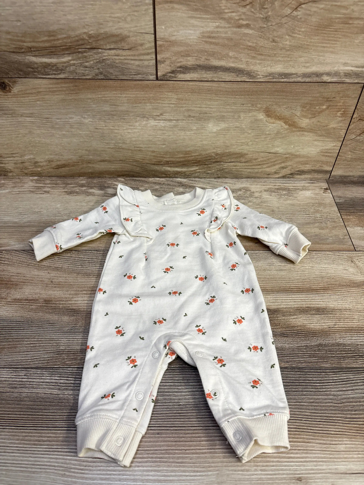 Front view of Carter’s ivory coverall with small pink floral print, ruffle trim, and snap leg closures on wood