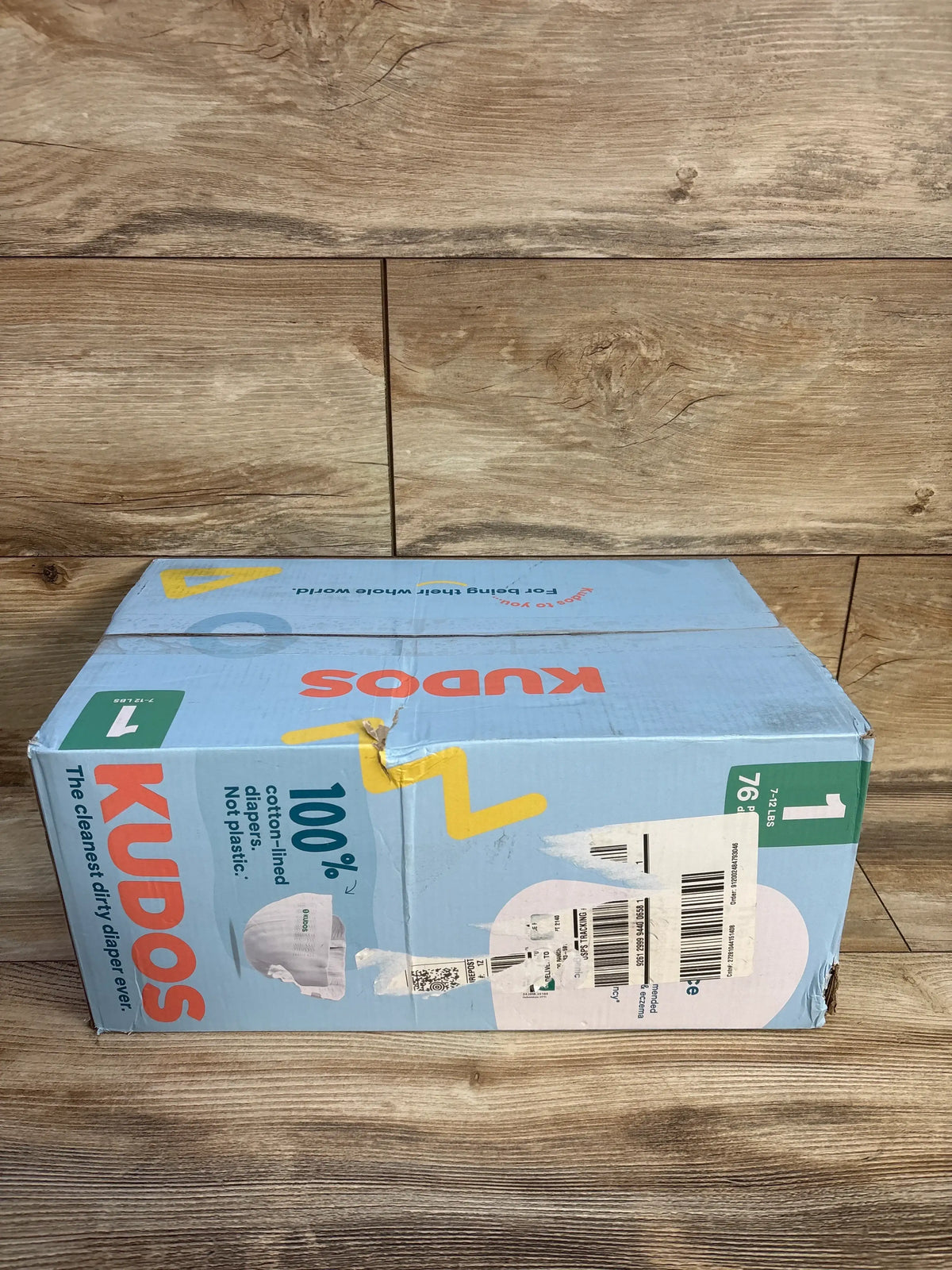 Angled view of Kudos size 1 disposable diaper box showcasing 100% cotton lining and 76 diapers on wood flooring