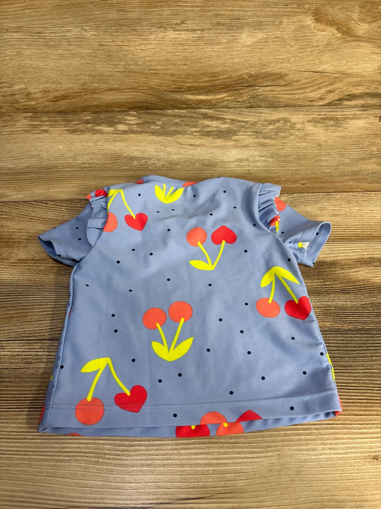 Back view of Carter’s blue cherry-print rashguard shirt (24m) showcasing ruffled short sleeves and bright cherry pattern on wood background
