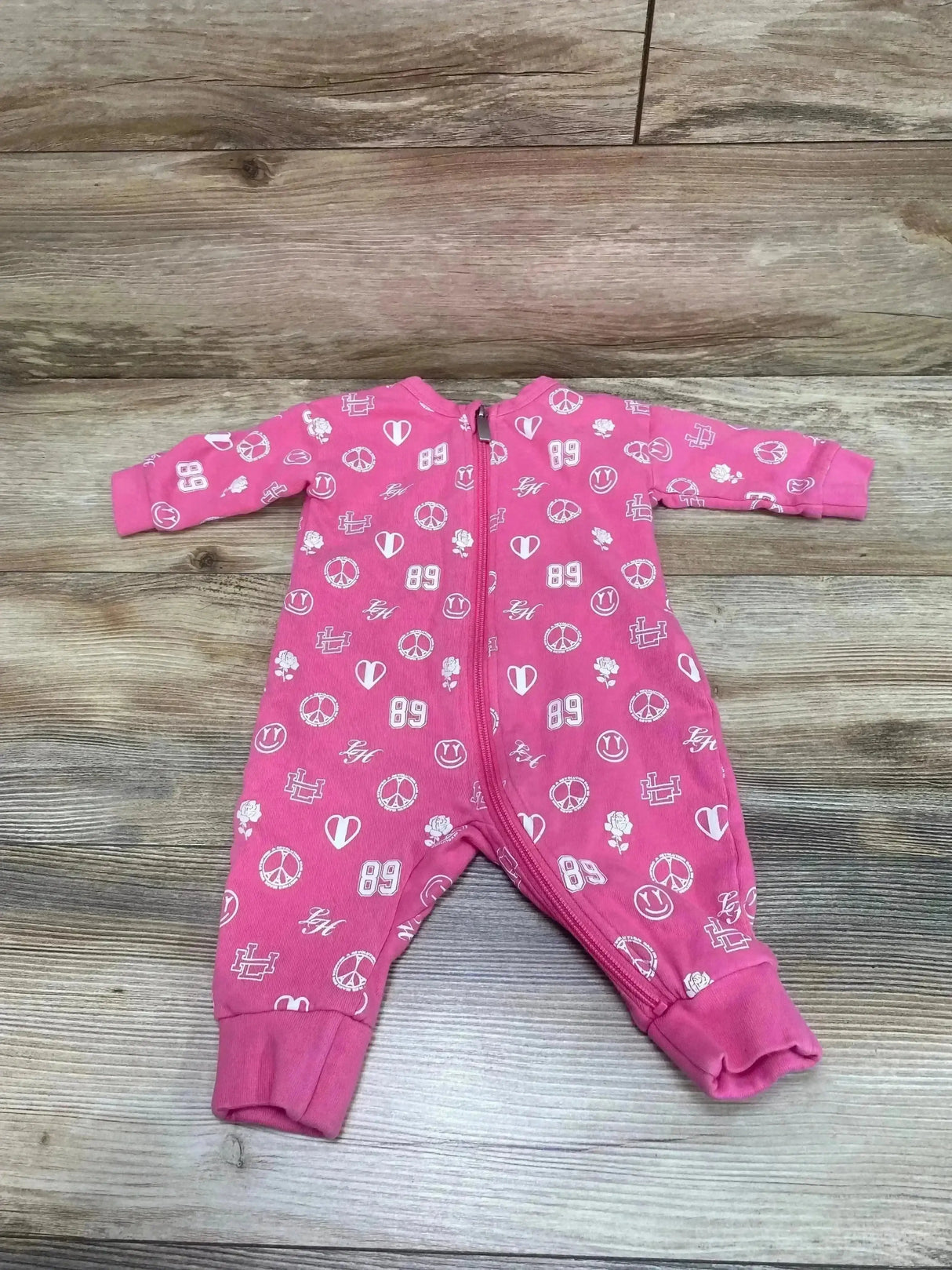 Front view of pink baby romper with white peace sign, heart and ‘89’ prints, front zipper and cuffed legs, laid flat on wood background
