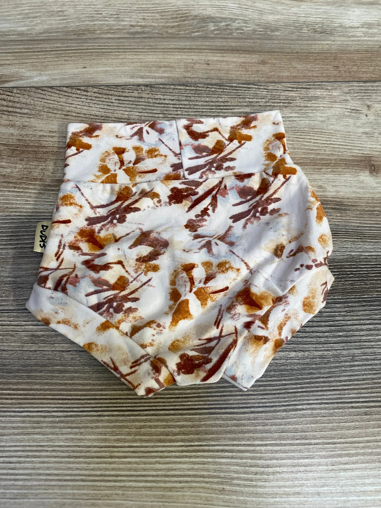 Top-down view of Dudis beige bummie (size 3-6m) showing rust and mustard floral print on white knit fabric against wood-grain background