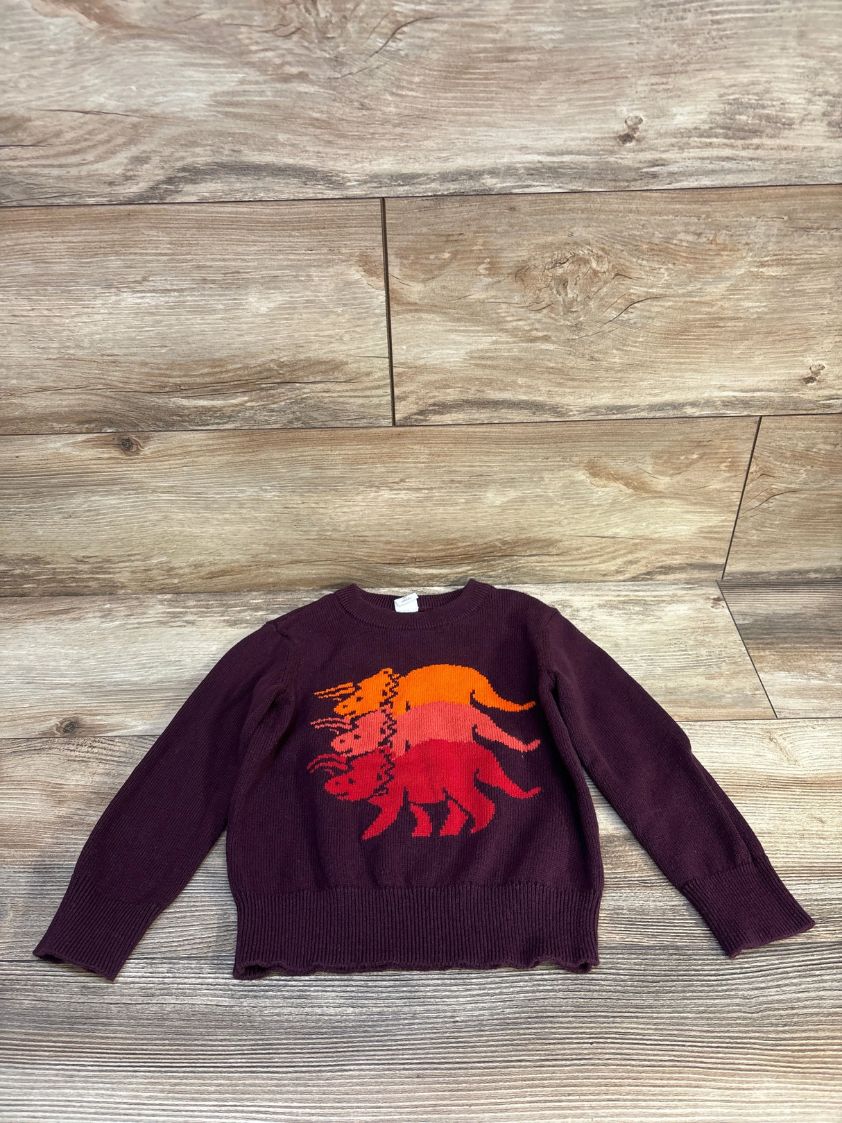 Burgundy kids sweater with colorful dinosaur graphic, long sleeves, laid on wood floor