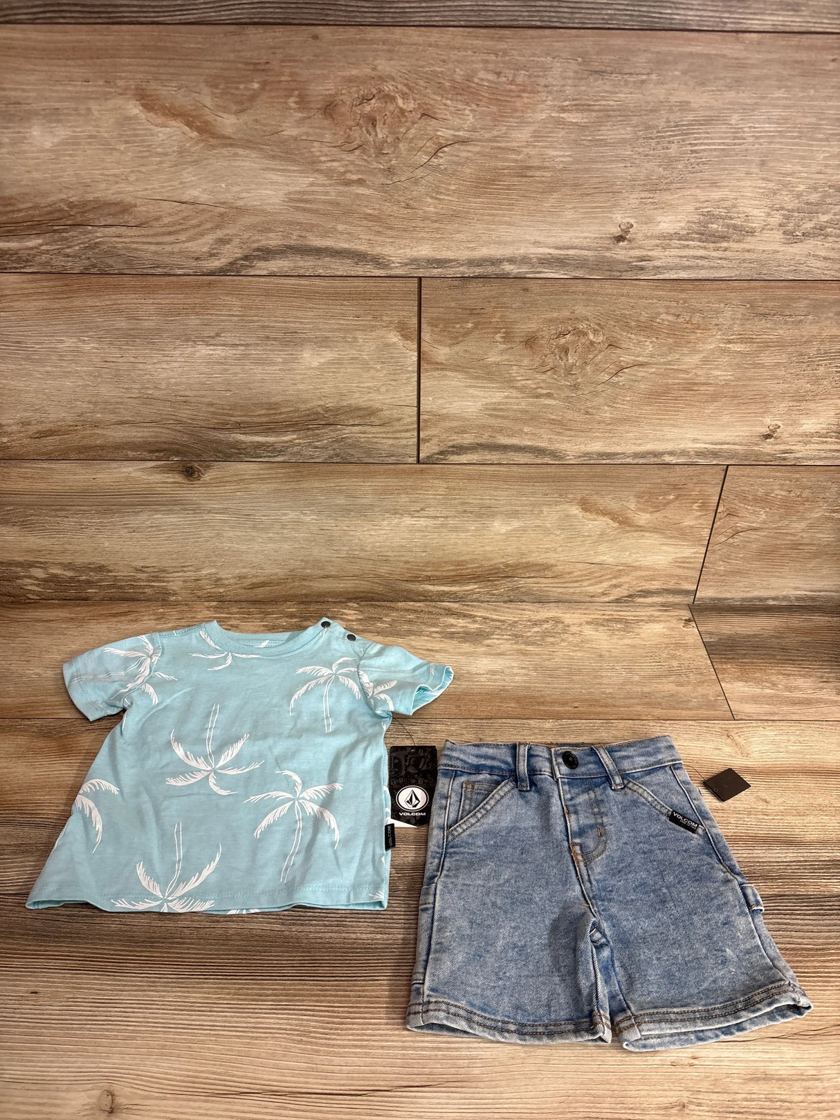 Flat lay of Volcom two-piece baby outfit showing light blue palm-tree T-shirt and jean shorts on wood floor
