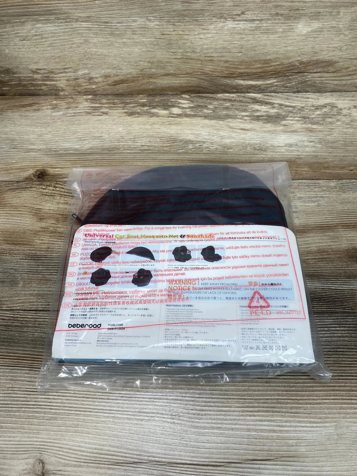 Top-down view of black Beberoad Love universal car seat mosquito net and sunshade folded in clear plastic packaging on wood tabletop
