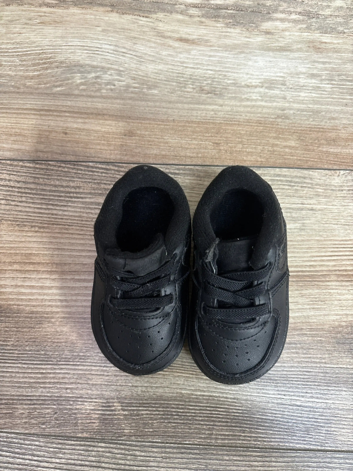 Top-down view of Nike Air Force 1 crib shoes, black size 4C, showcasing perforated toe, laces, and padded collar on wood floor