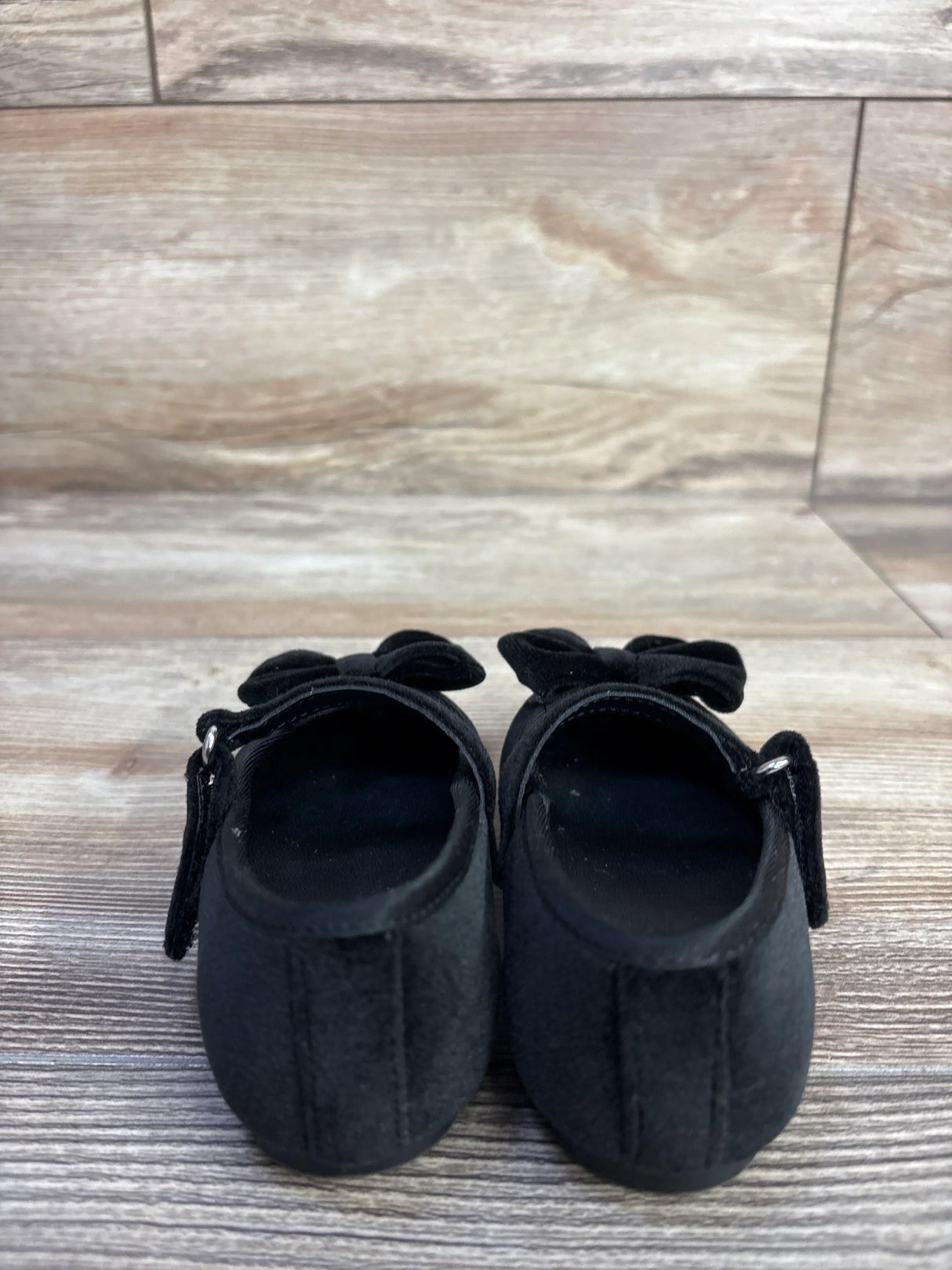 Rear view of Baby Gap velvet Mary Jane flats, black size 5C, showing bows and Velcro straps on light wood floor background