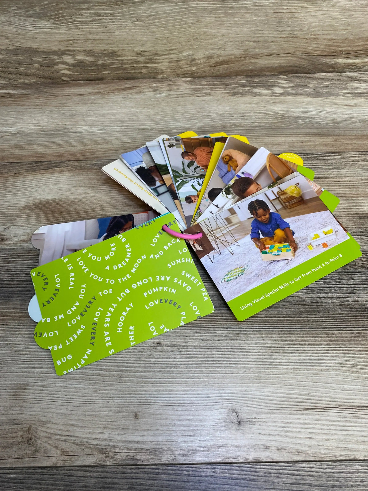 Angled view of Lovevery Persister Play Guide fanned to show ring-bound tabbed activity cards with green cover on wood table