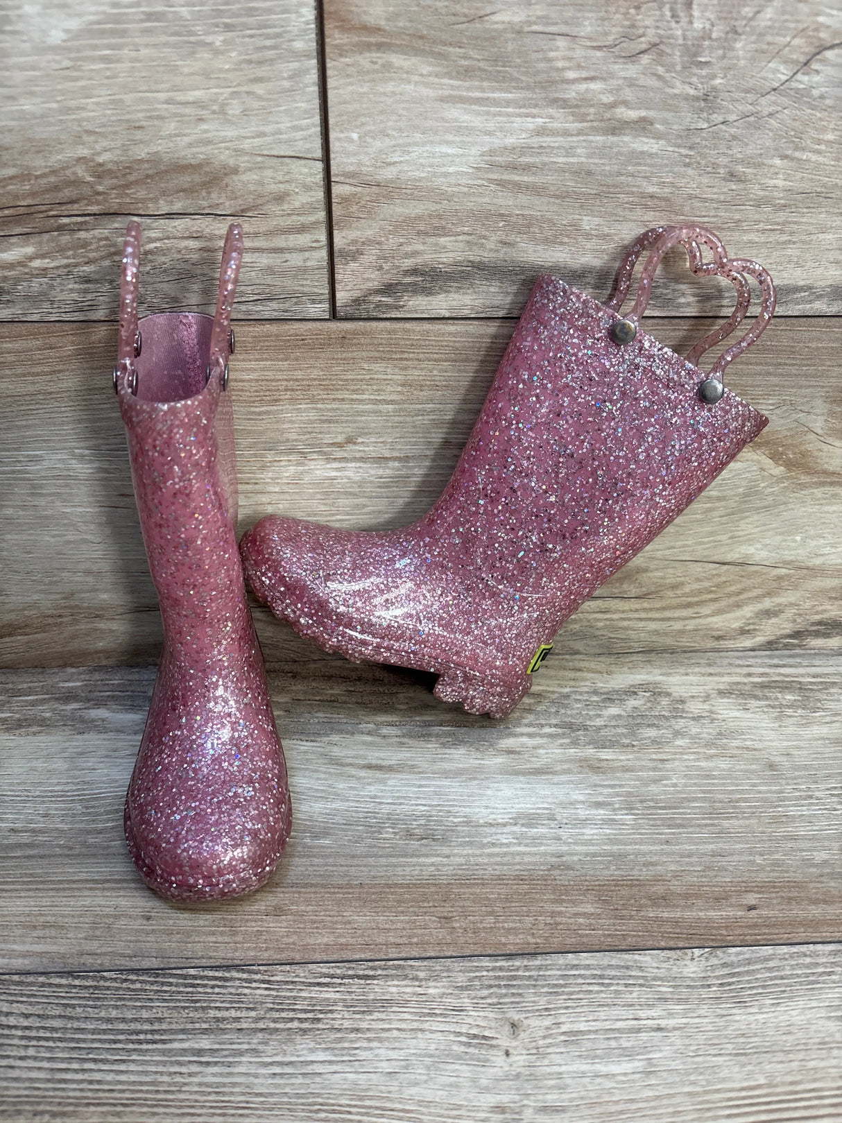 Pink glitter kids rain boots with heart-shaped handles on wood background