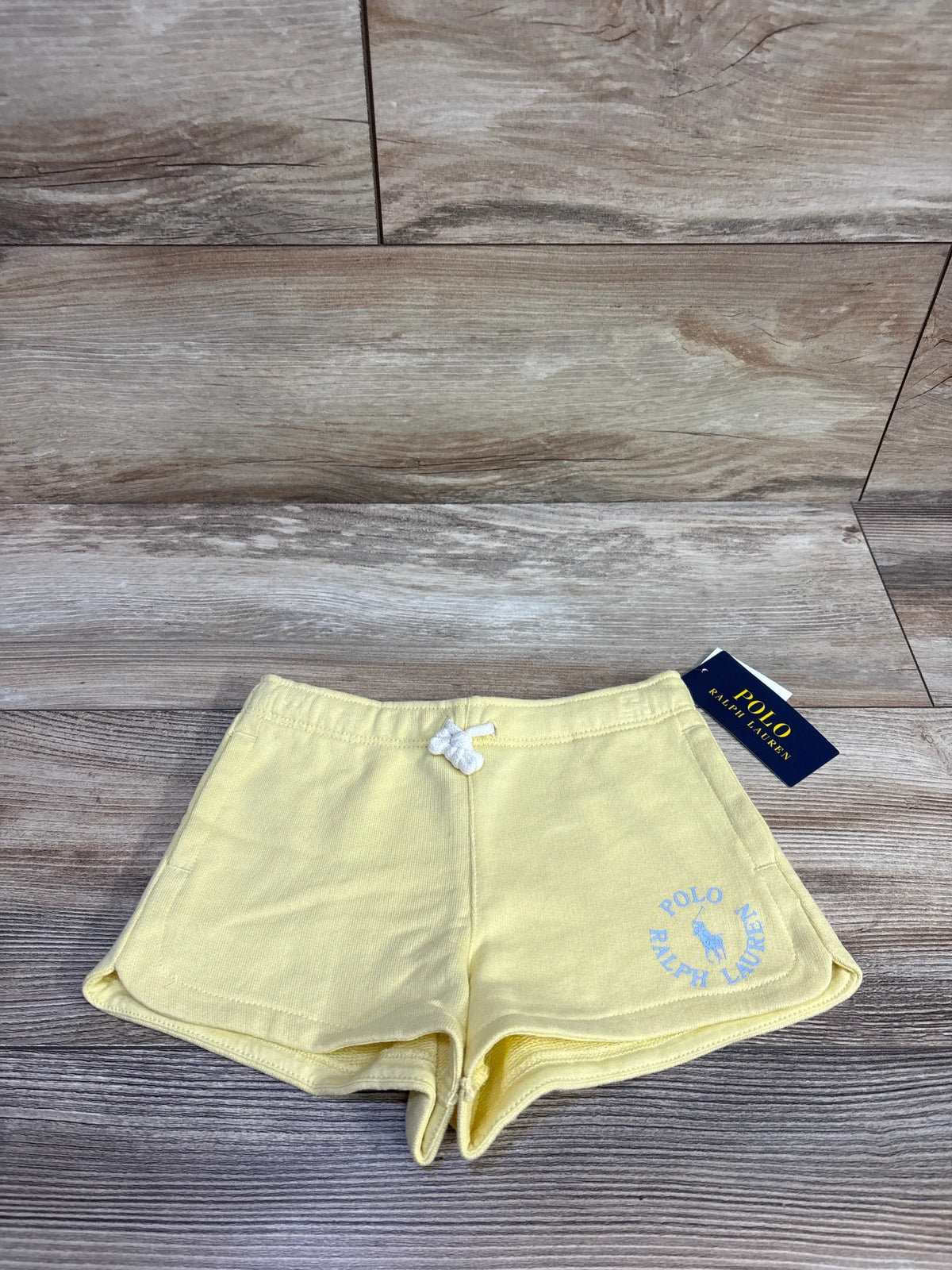Front view of Longwood Yellow Polo Ralph Lauren drawstring shorts showcasing a blue logo and scalloped hems on wood grain