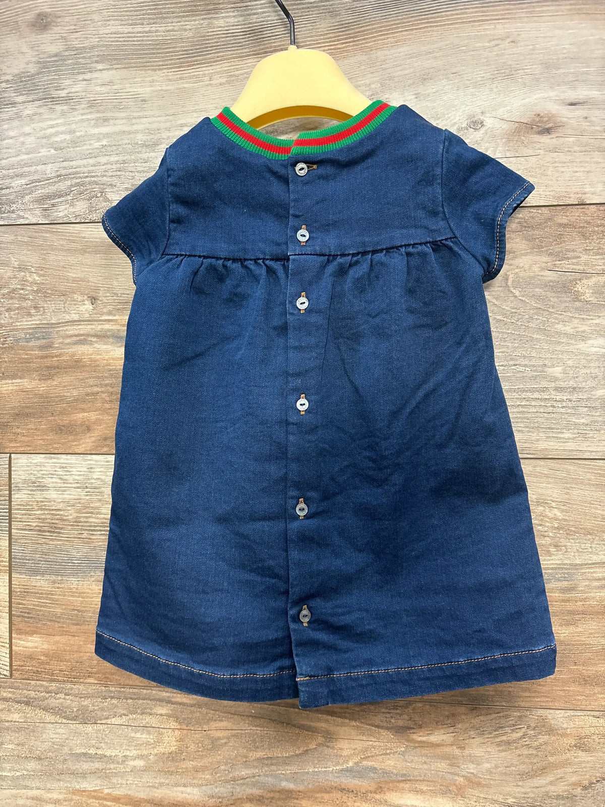 Back view of Gucci blue denim baby dress with red-green ribbed collar and back button closure on yellow hanger over wood floor