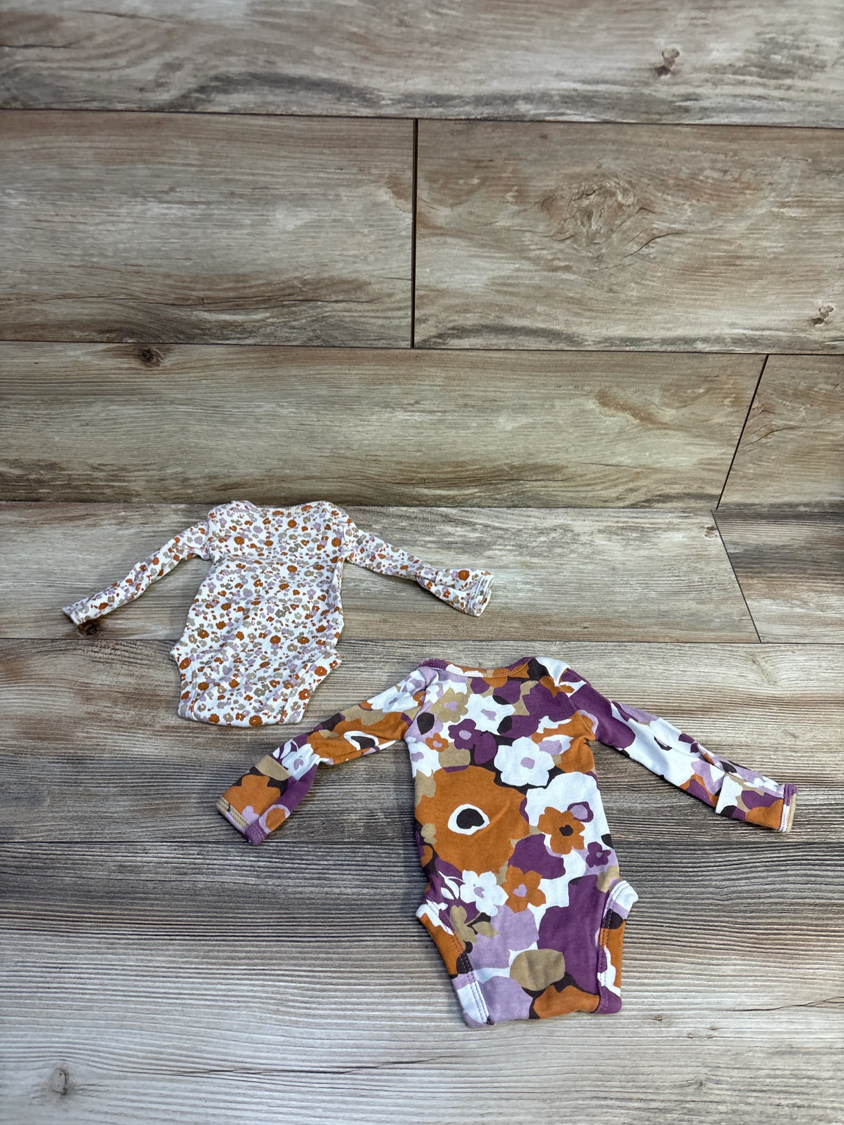 Flat lay of two Carter’s preemie long-sleeve bodysuits showcasing orange, purple, and brown floral prints on wood background