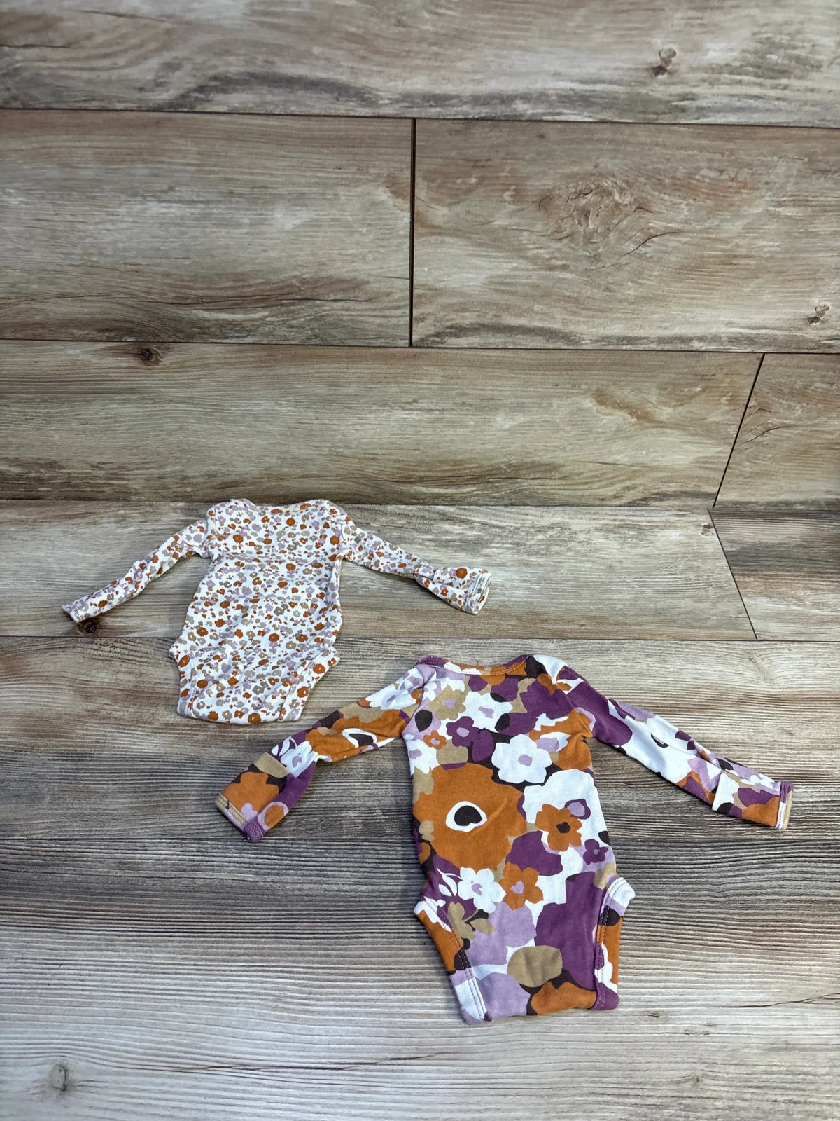 Flat lay of two Carter’s preemie long-sleeve bodysuits showcasing orange, purple, and brown floral prints on wood background