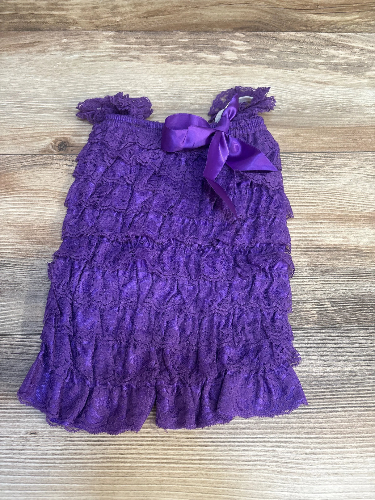 Purple lace ruffle baby romper with satin bow, displayed on wood background.