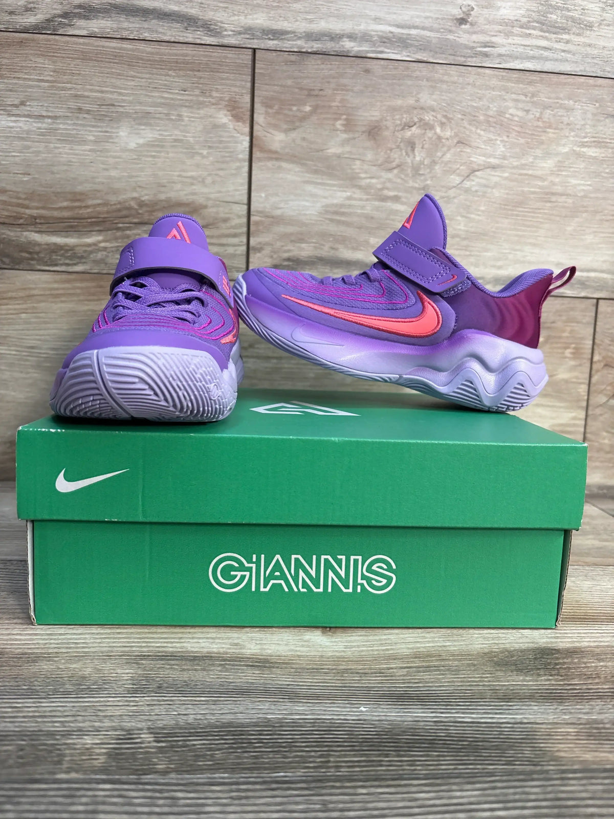 Purple and pink Nike Giannis kids sneakers on green shoe box, wood background
