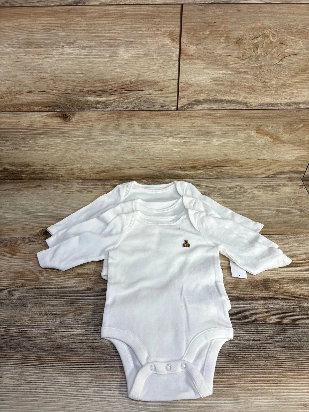 Top-down view of three white Baby Gap long-sleeve embroidered bodysuits stacked, showing small brown teddy bear embroidery on chest, wood plank background