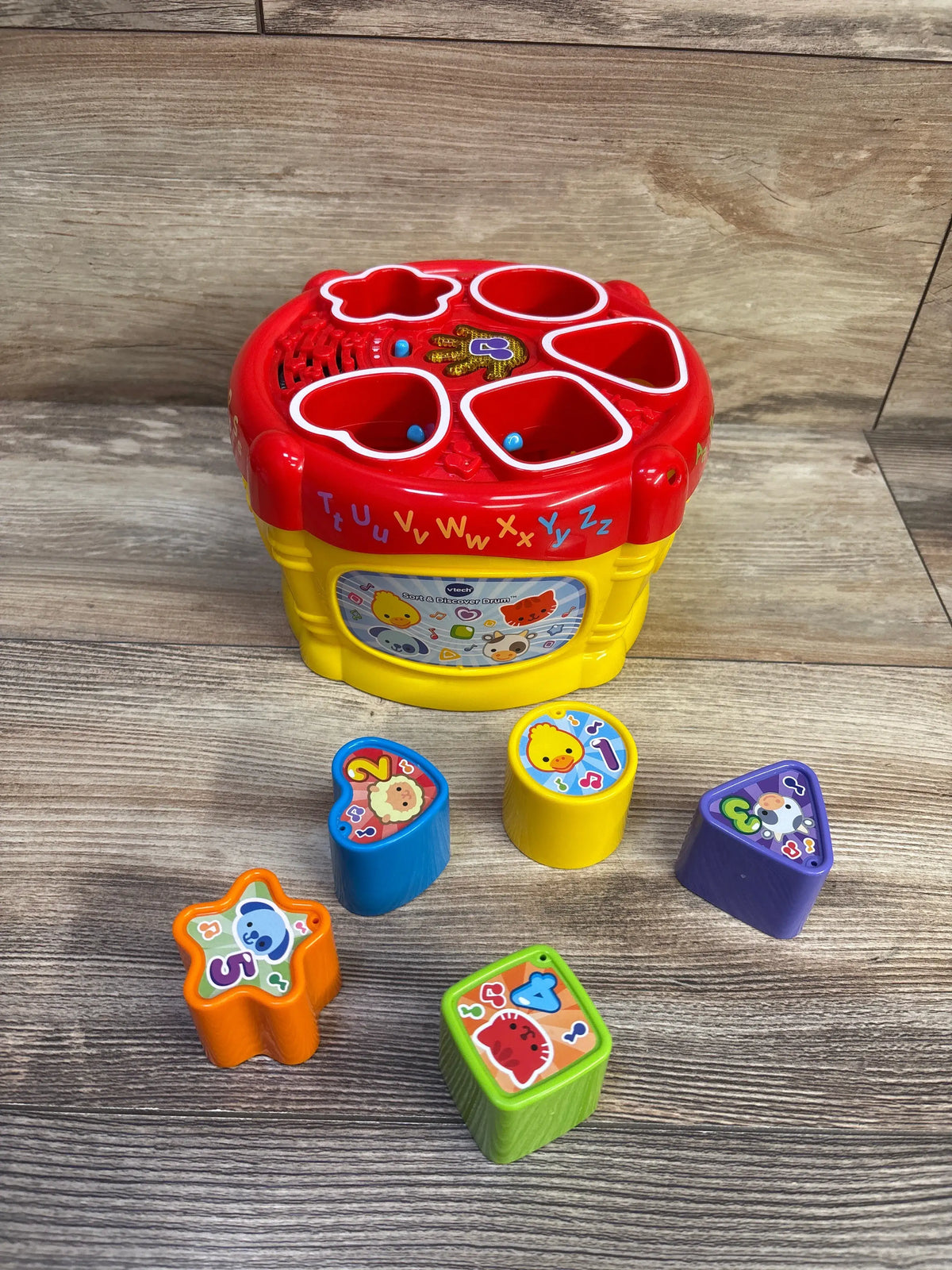 Angled view of VTech Sort and Discover Drum showing red lid with five shape slots and six colorful blocks on wood background
