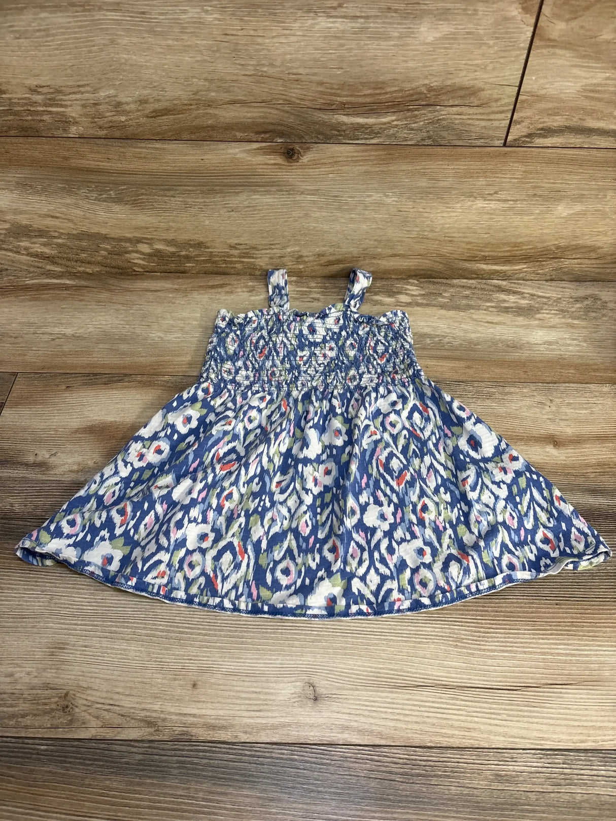 Flat-lay of blue Oshkosh smocked 3T toddler dress with smocked bodice, thin straps, and floral ikat print on wood floor