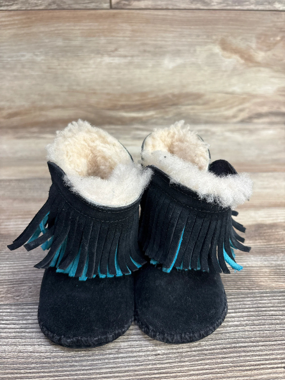 Black baby moccasin boots with cream fleece lining and turquoise-accented fringe on wooden floor