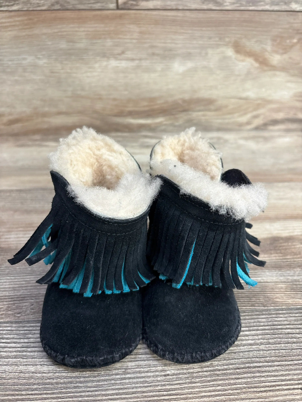 Black baby moccasin boots with cream fleece lining and turquoise-accented fringe on wooden floor