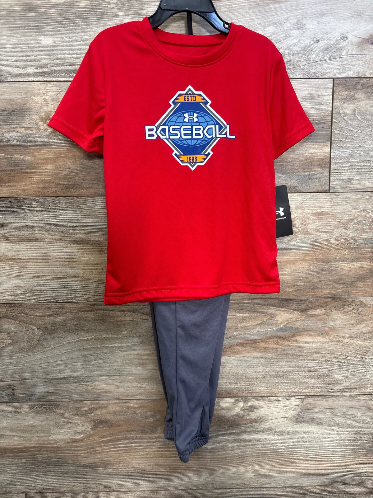 Front view of Under Armour red toddler shirt with large Baseball logo and gray elastic hem pants on wood background