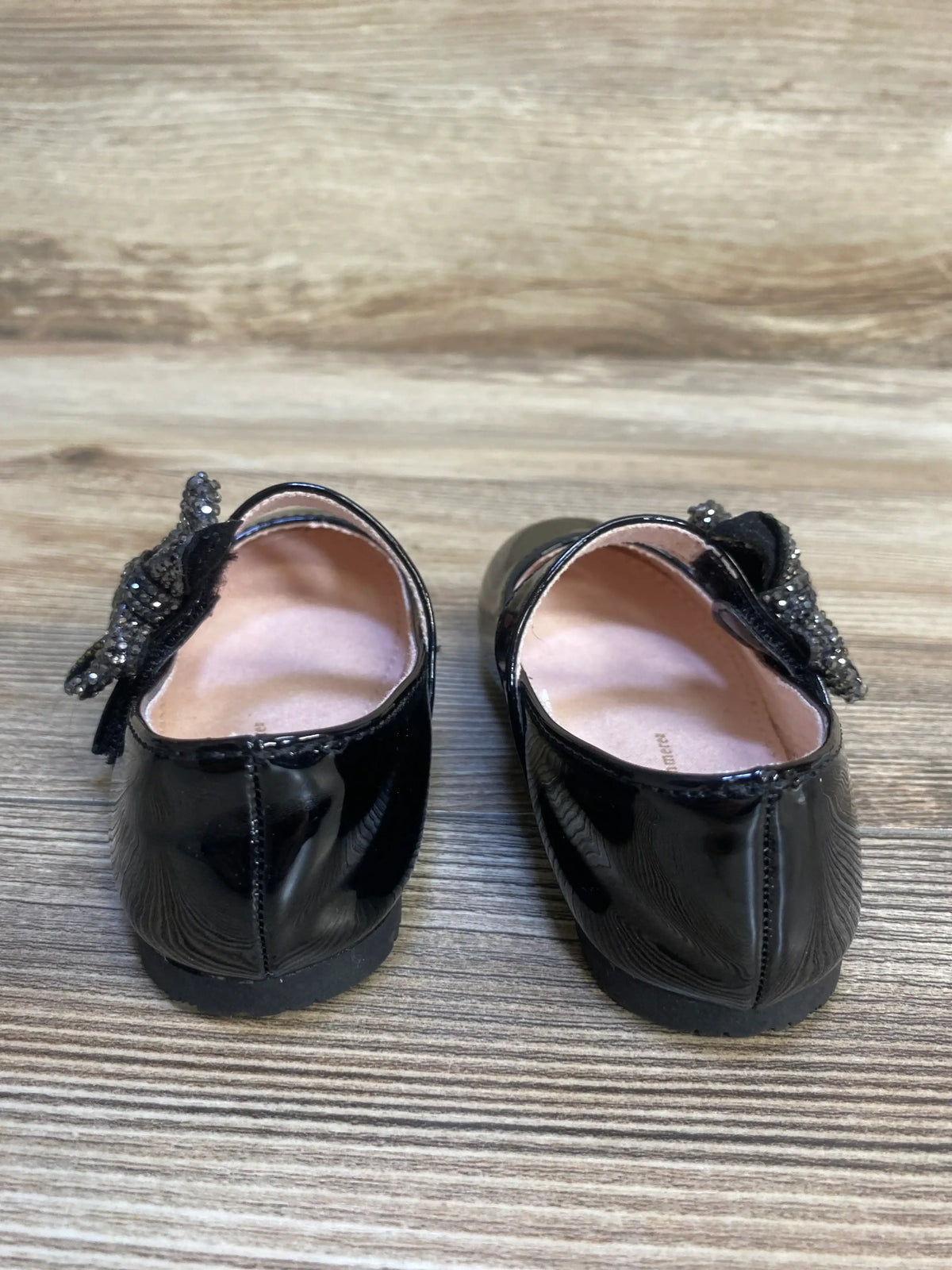 Rear view of Cupcakes & Cashmere black patent Mary Jane flats, size 6C, showing sparkly beaded bows on wood floor