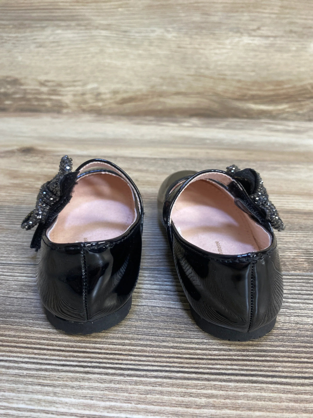 Black patent leather girls dress shoes with sparkly bow detail, displayed on wood floor