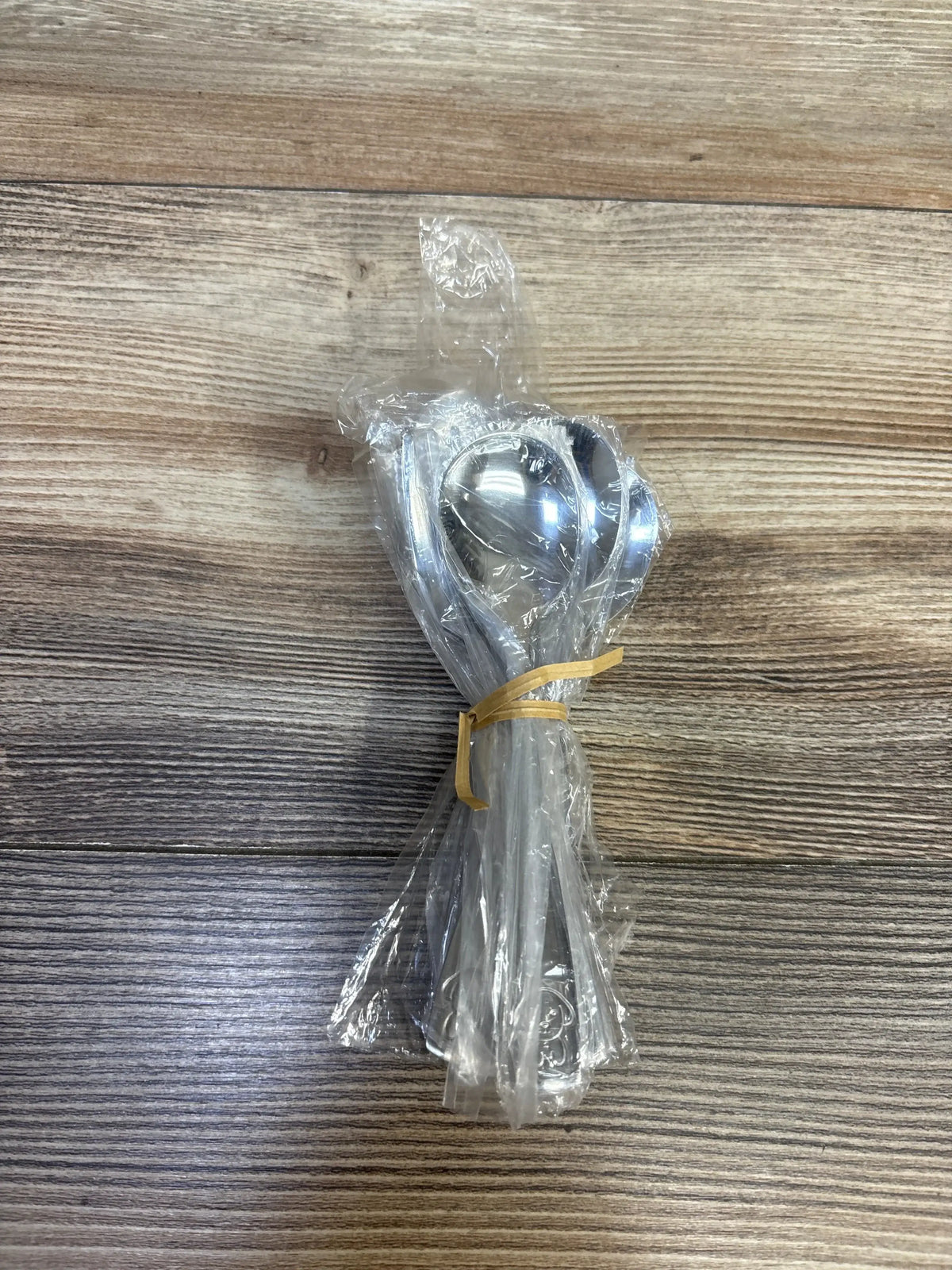 Overhead view of 10pc stainless steel toddler utensils in clear plastic tied with a rubber band on wood surface