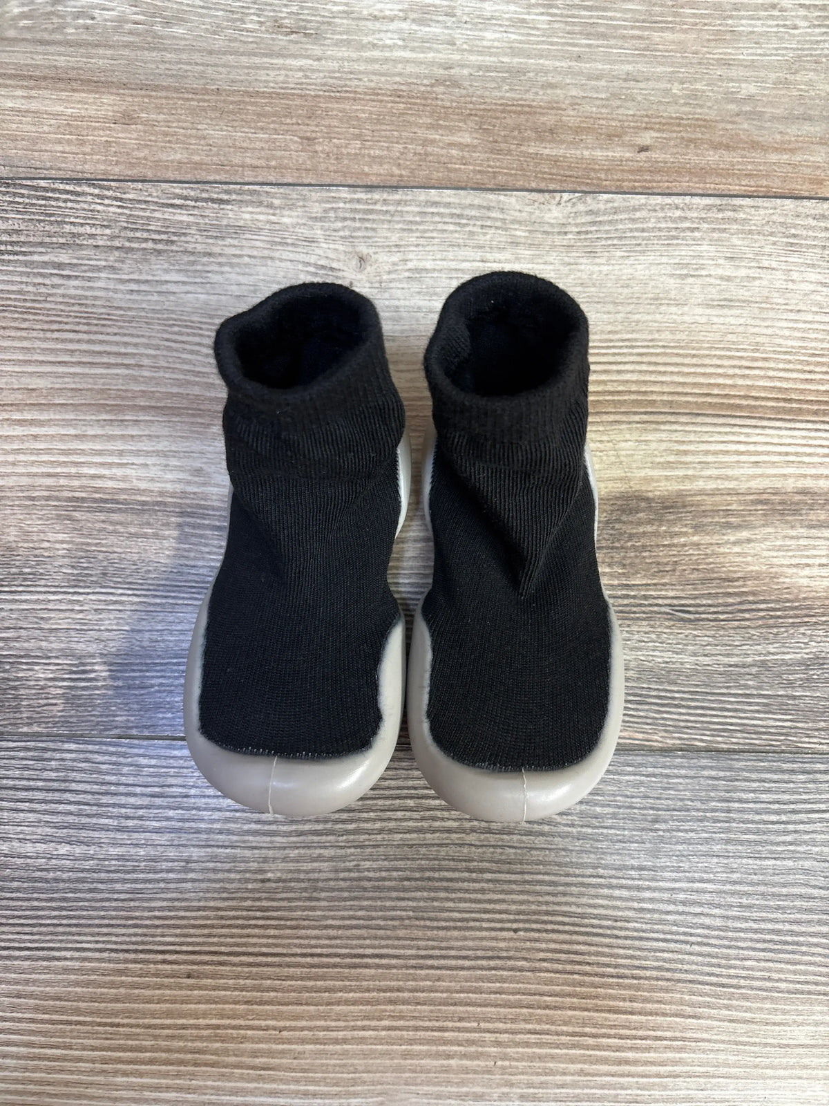 Top view of black baby sock shoes showcasing soft rubber soles on a wood-grain surface