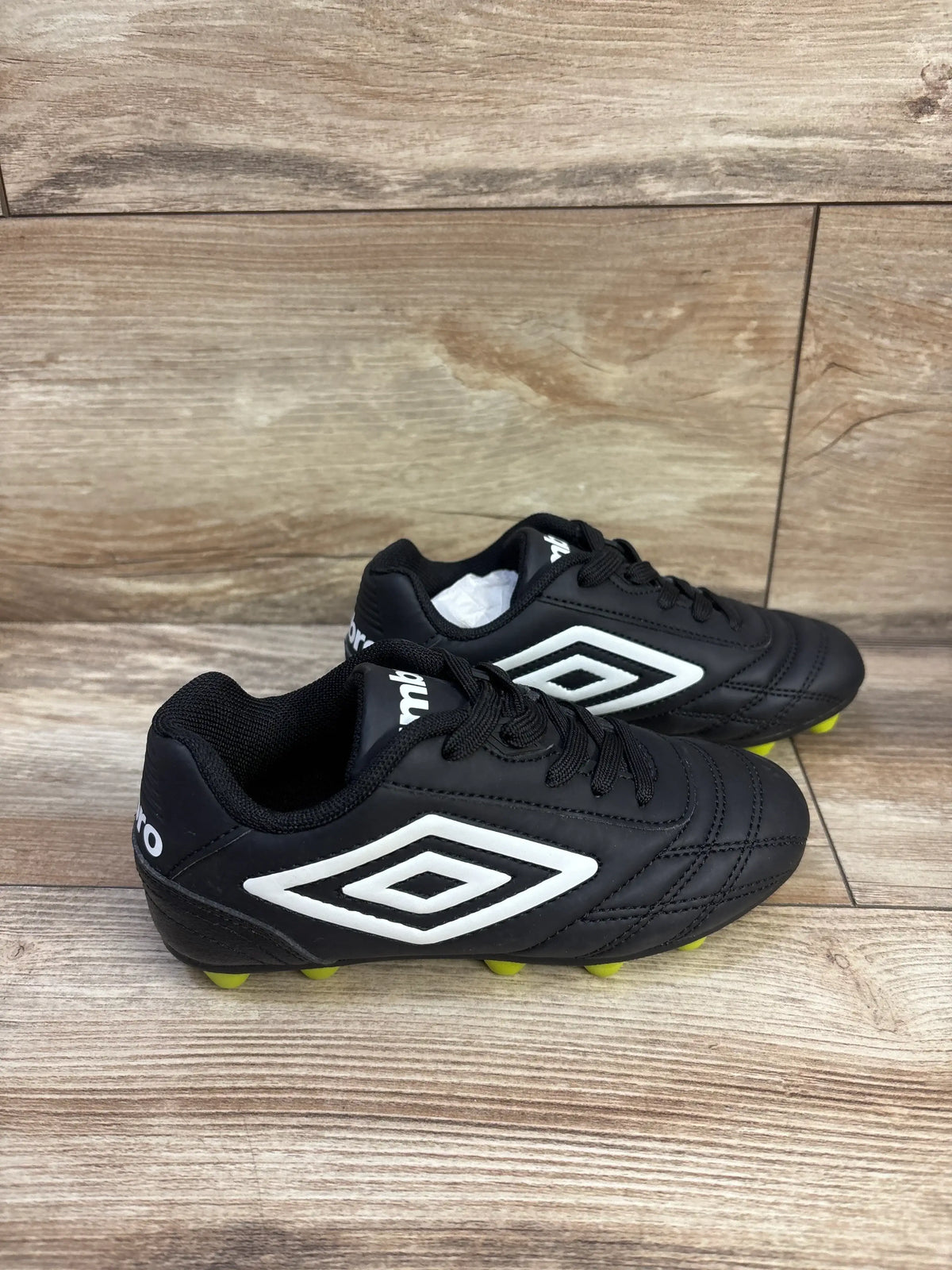 Side view of Umbro kids’ black soccer cleats size 11C showing white double-diamond logo and yellow studs on wood background