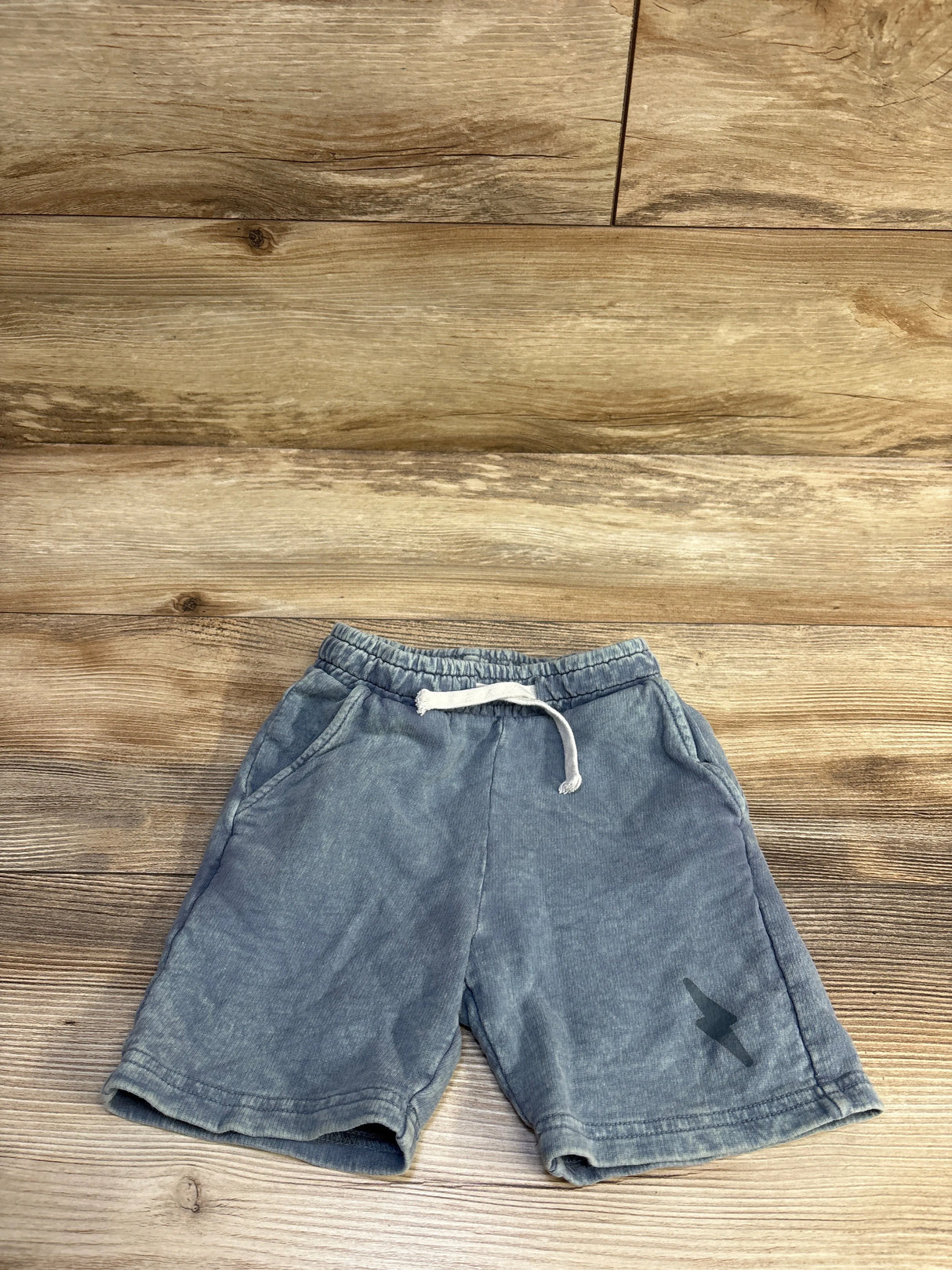 Flat-lay front of toddler blue drawstring shorts with elastic waistband and white drawstring, side pockets, faded wash and small lightning bolt on wood background