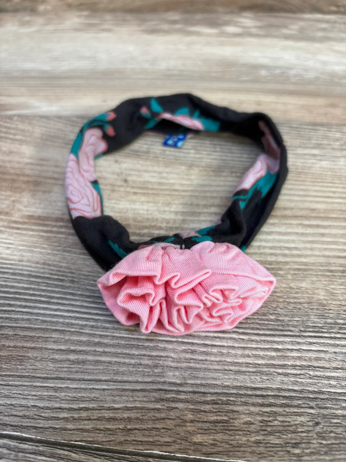 Close-up front view of Kickee Pants floral baby headband in black with pink ruffle flower detail on wood surface