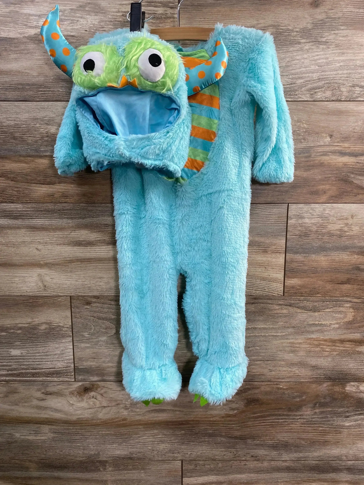 Front view of Spirit Lil’ Monster baby costume showcasing fuzzy turquoise one piece with monster-face hood, orange striped horns, and footed legs on wood background